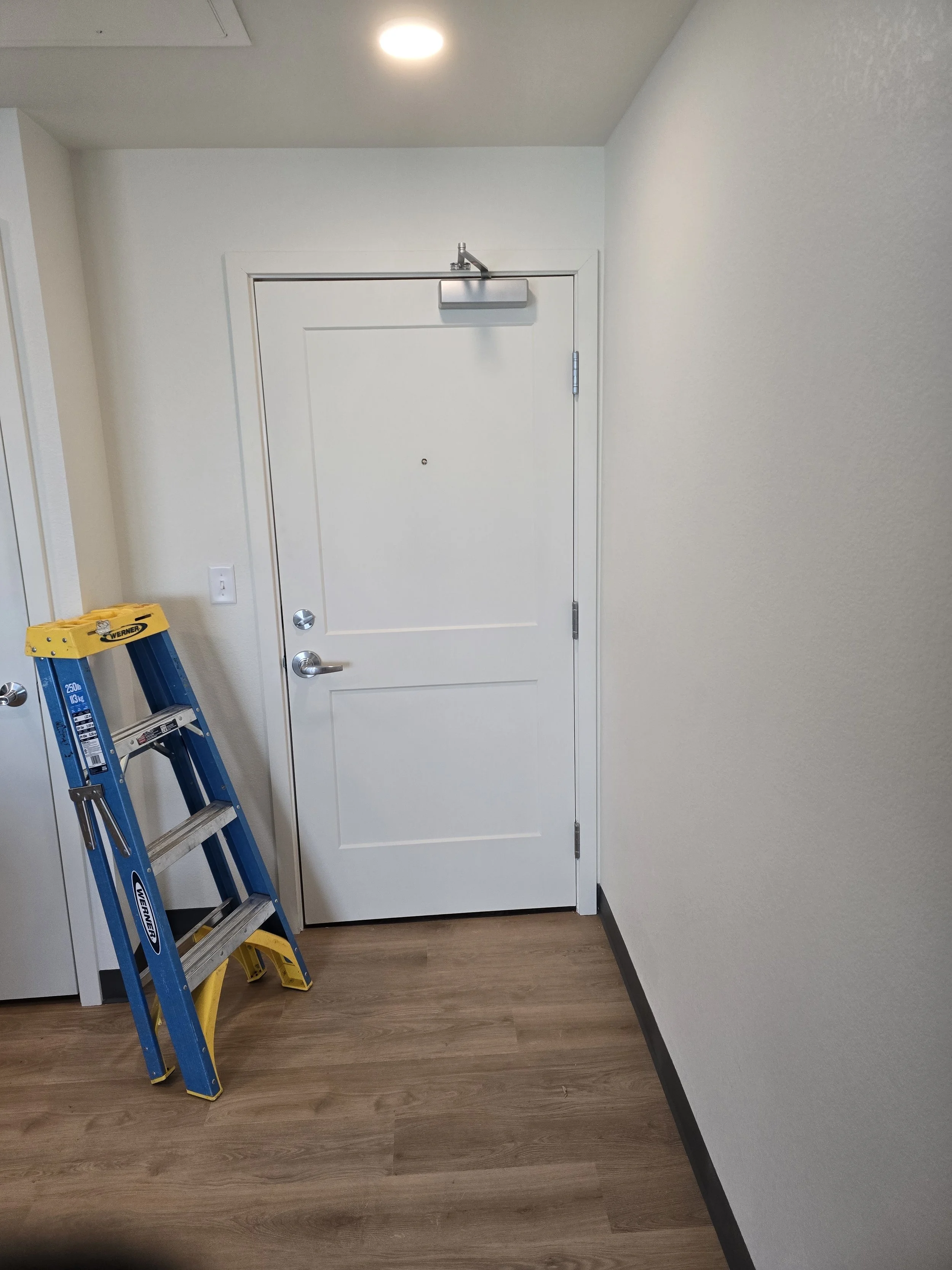 Interior view of an apartment entry door with a small peephole, accompanied by a blue and yellow step ladder leaning against the wall on the left.
