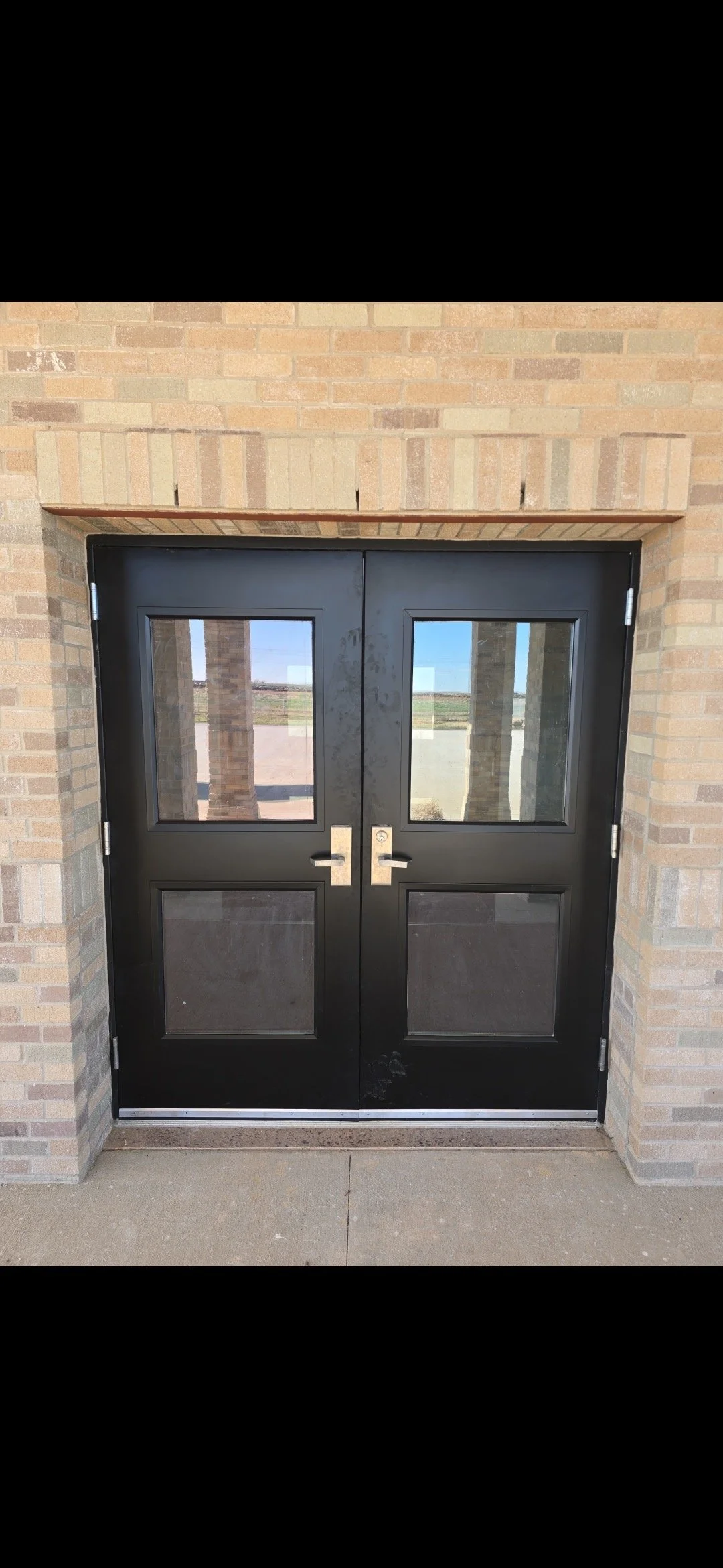 Black double doors with glass panels, set in a brick wall, with a concrete sidewalk in front.