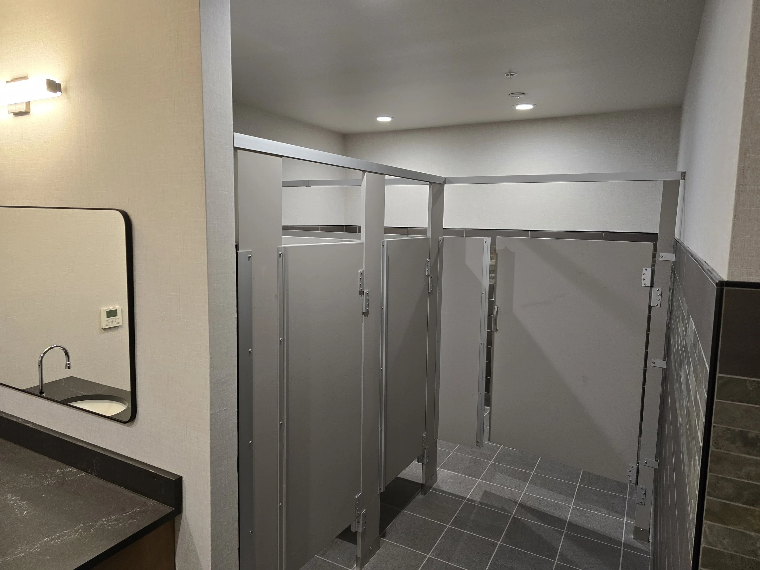 Public restroom with two gray stalls, a mirror on the left wall, black countertop with a sink, and beige walls.