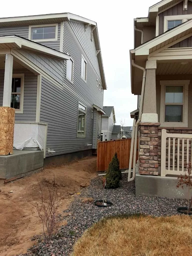 View of two houses with a narrow backyard, a small tree planted in gravel, and a wooden fence between the properties. One house has gray siding, and the other has beige siding with brick accents.