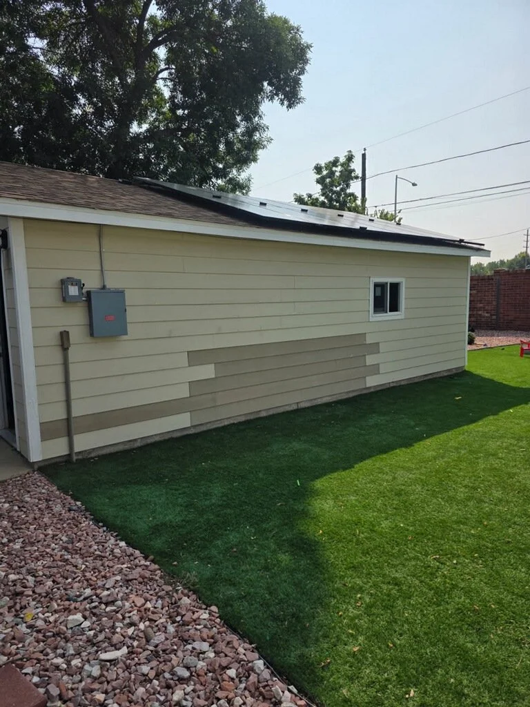 A backyard house with beige siding, small window, solar panels on the roof, and a grassy lawn with a gravel border.