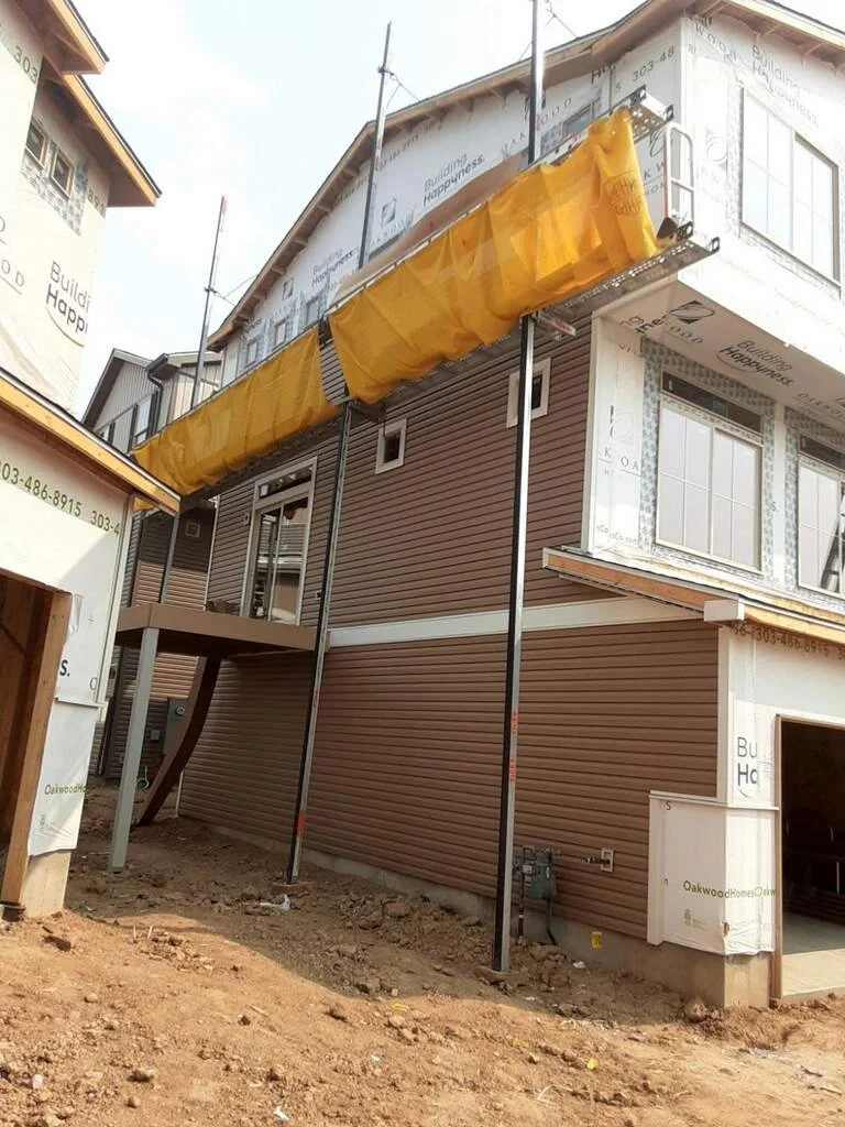 Under construction house with scaffolding and yellow protective cover, brown siding, and exposed ground in front.