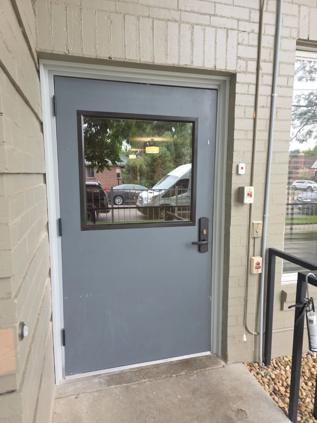 Gray door with a window reflecting trees and parked cars outside, with a black handle and lock, in a beige brick building.