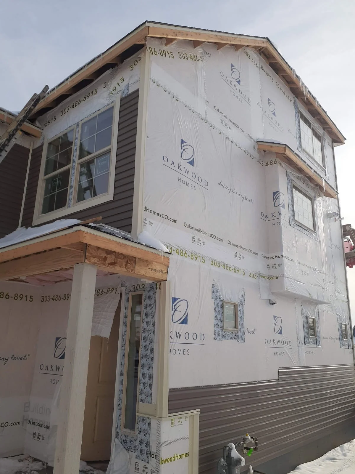 A multi-story house under construction wrapped in white weather-resistant building paper with the logo 'Oakwood Homes'. The house has several windows, some installed and others not yet with finished trim. There is a small covered porch area supported
