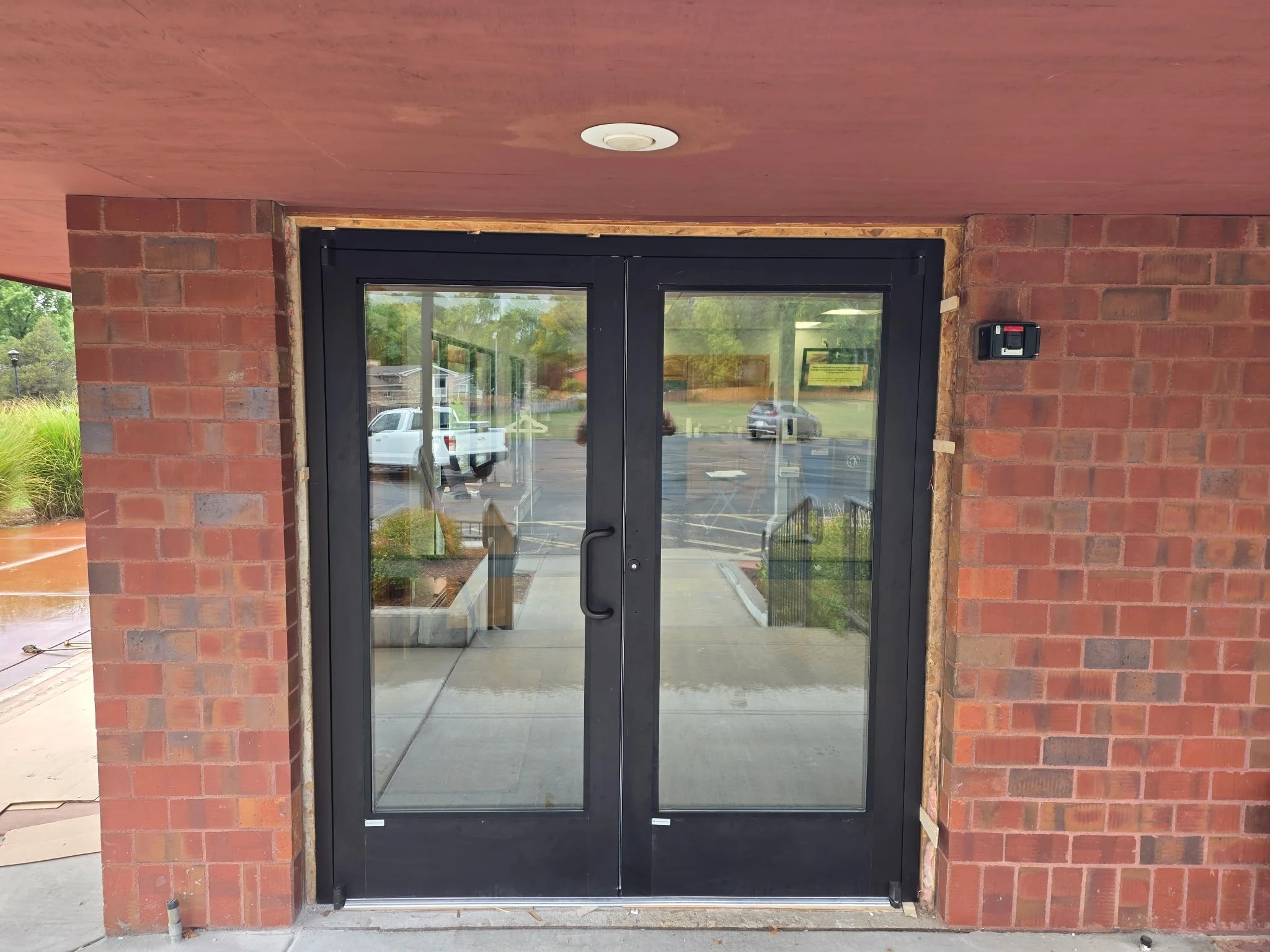 Entrance with black-framed glass double doors, brick exterior, and an access control keypad on the right wall.