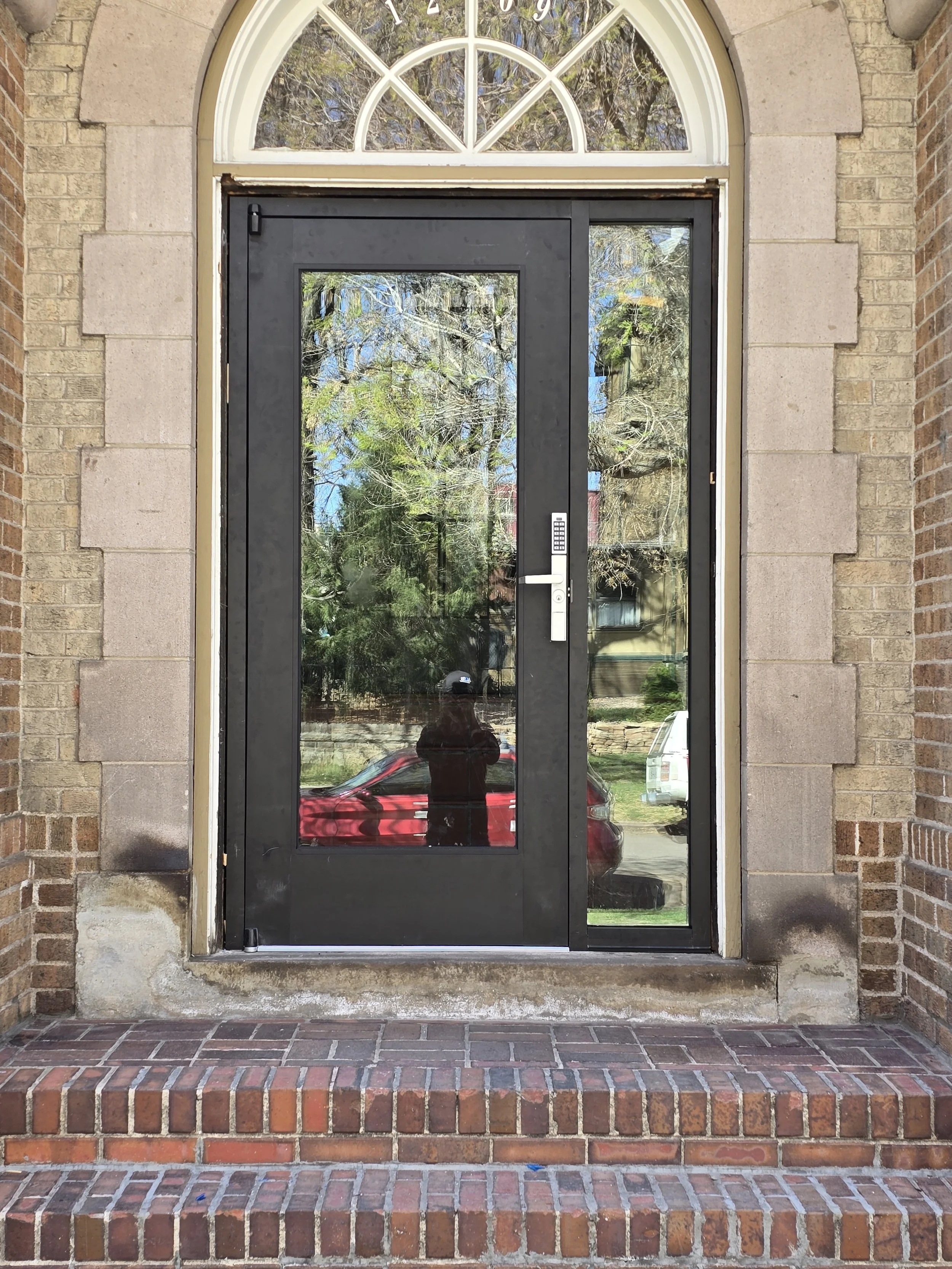 Black front door with glass panels and a digital keypad, framed by brick and stone, with a semicircular window above showing reflections of trees and sky.