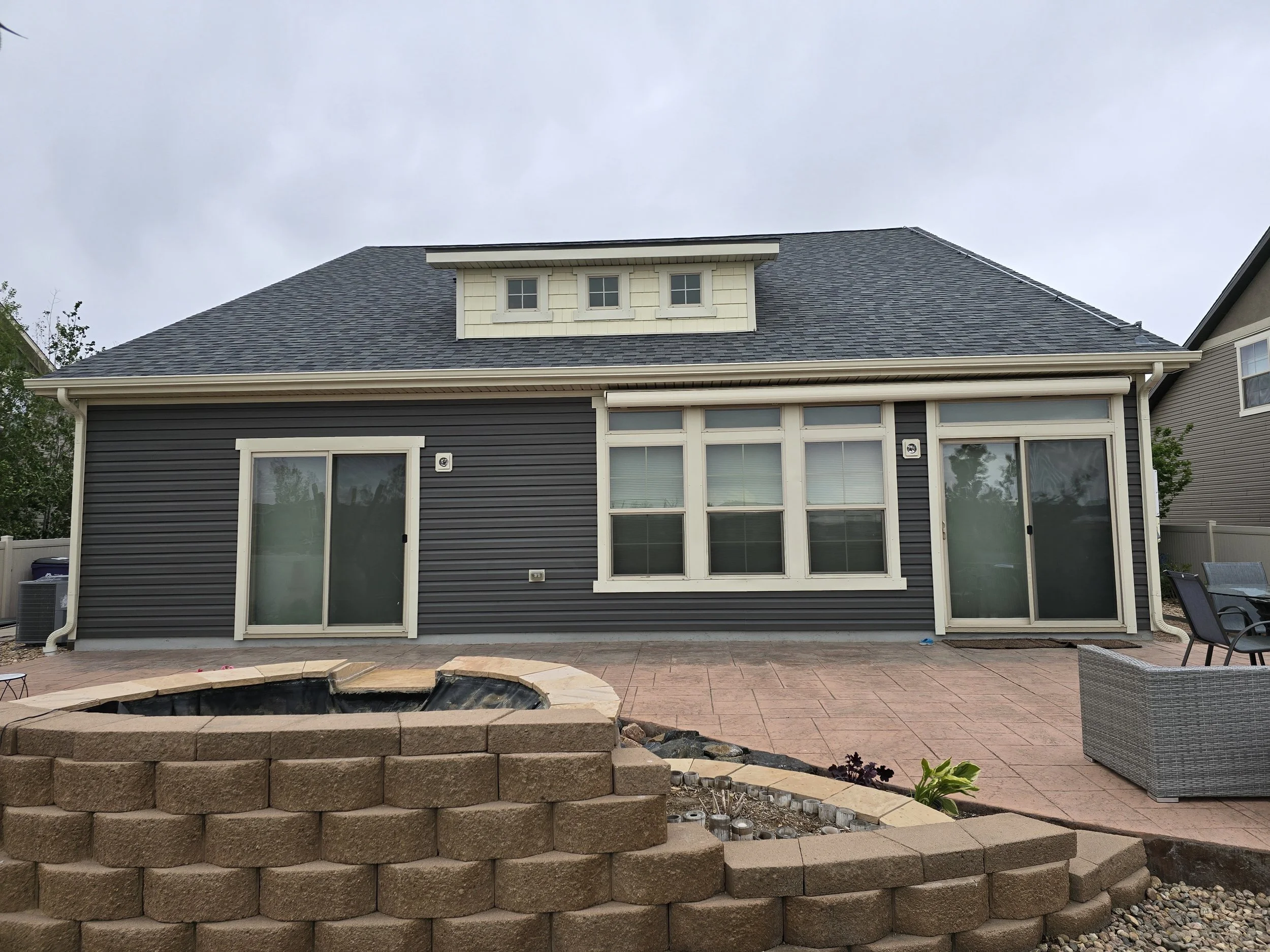 Backyard with a house featuring dark gray siding, sliding glass doors, and a small windowed dormer on the roof. The backyard has a brick patio, a fire pit, some outdoor furniture, and a small garden bed.