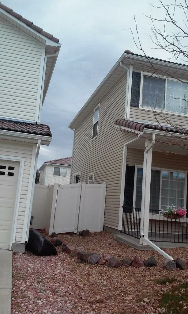 View of two houses with beige and white siding, a white fence, and a gravel yard with rocks. Overcast sky and leafless tree branches
