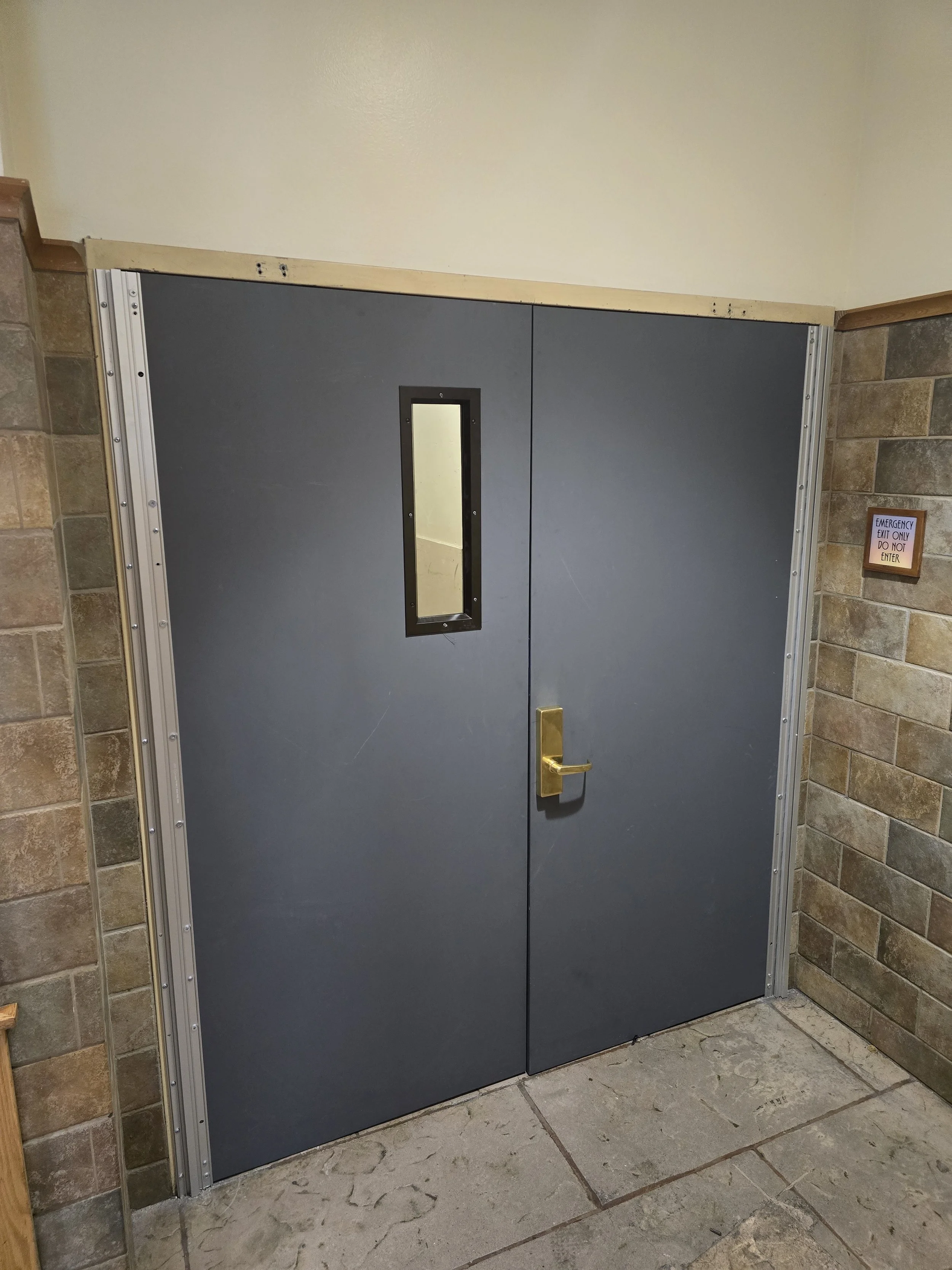 A blue double door with a small rectangular window and a golden handle, set in a brick wall with a handicap accessible door frame and a small sign on the wall.