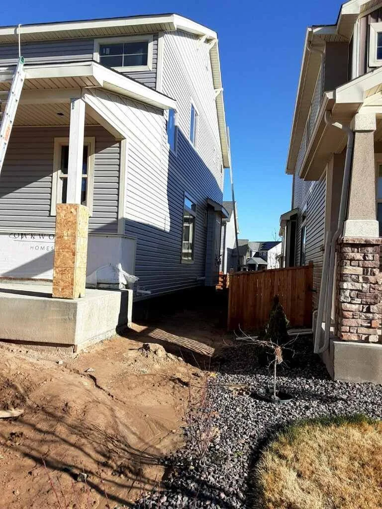 View of two houses with a gap between them. The house on the left is under construction with the foundation and some walls visible, while the house on the right is finished with a brick and siding exterior. A small new tree is planted in the yard, an
