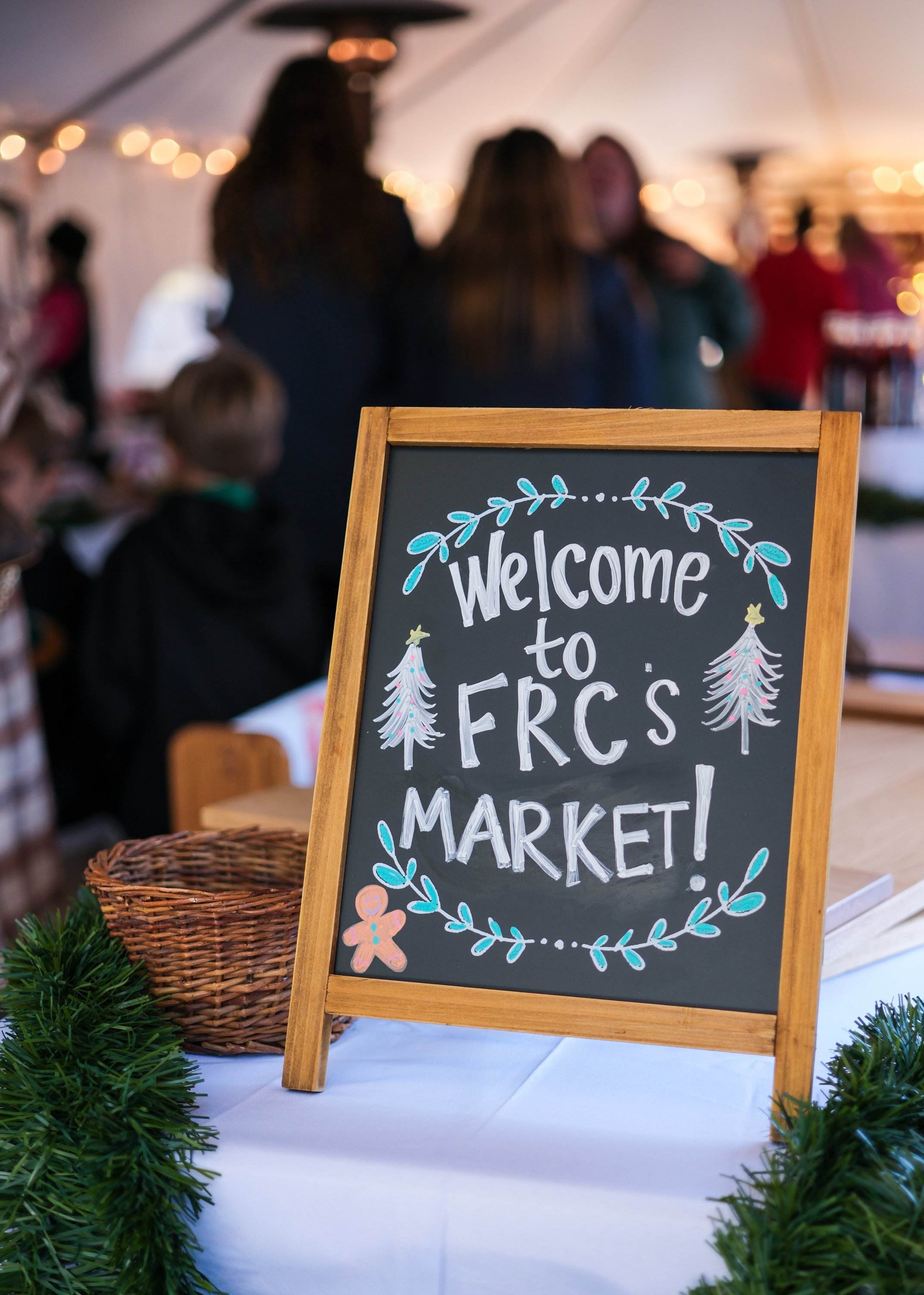 A chalkboard sign that says "Welcome to FRC's Market!" decorated with drawings of Christmas trees, leaves, and a gingerbread man, placed on a table with green garland in front. Blurred people are in the background at an indoor event.