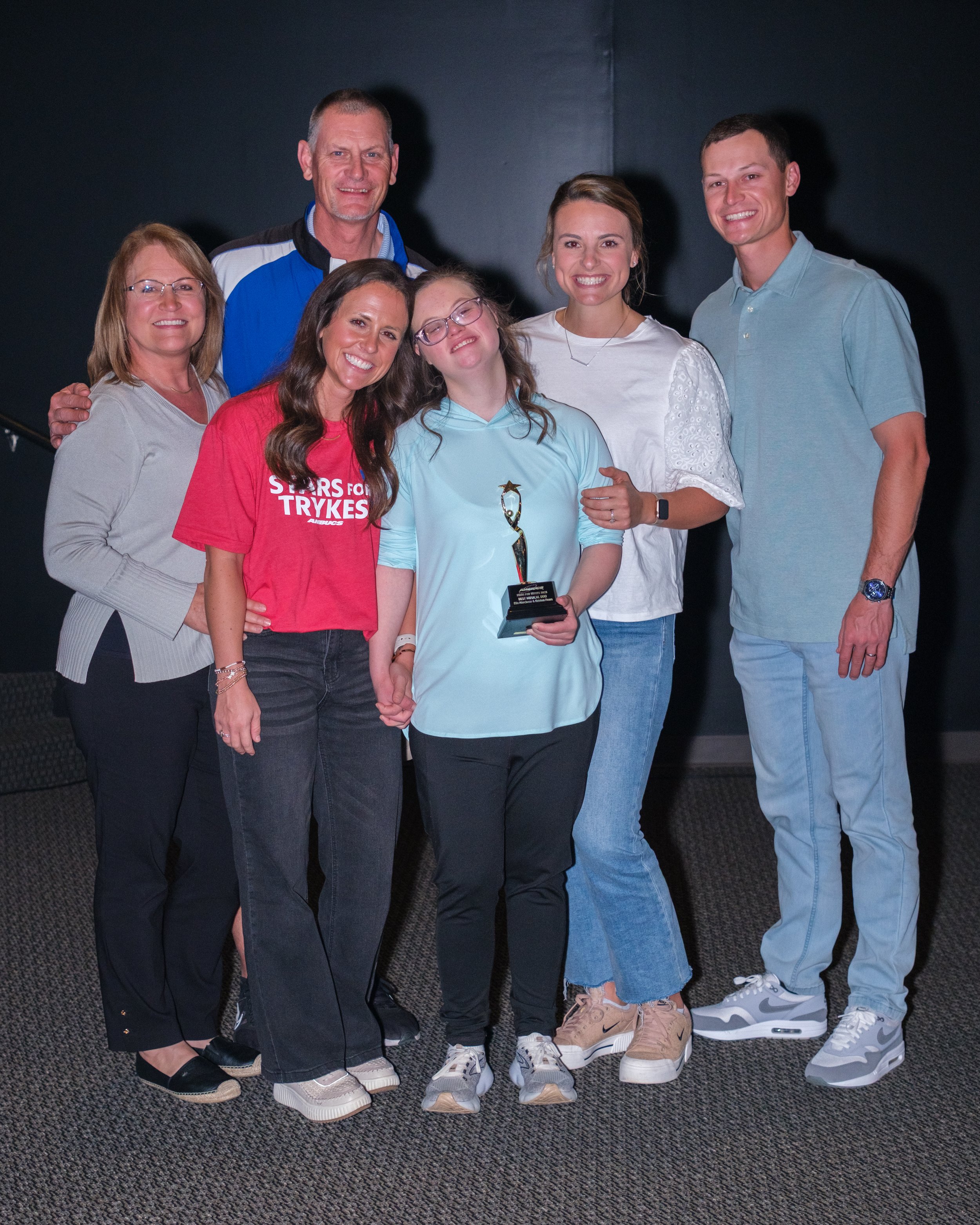 Group of six people posing together with a young girl holding a trophy, celebrating an achievement, on a black background.