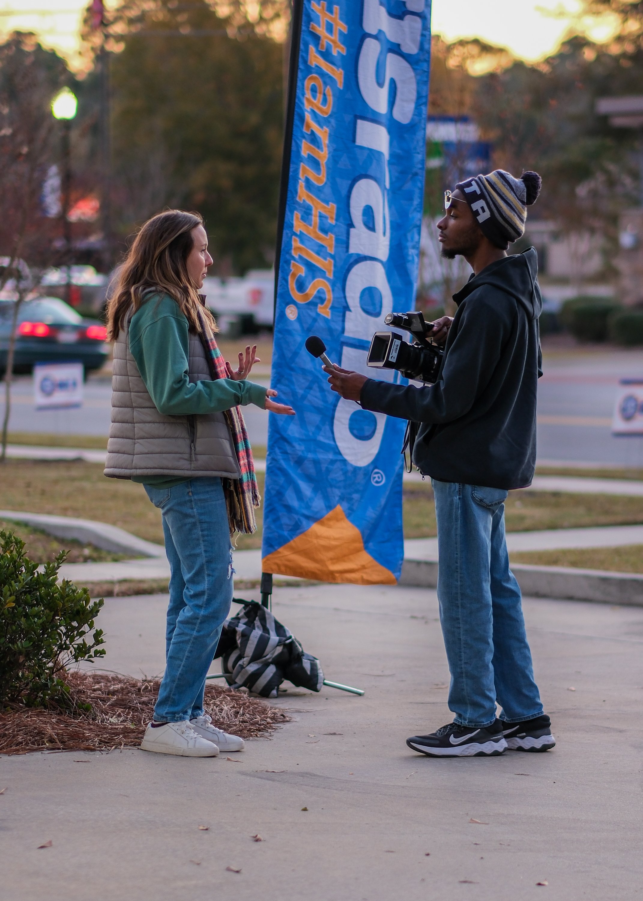 A woman being interviewed by a man with a video camera in an outdoor setting, next to a large blue banner with orange and white text.
