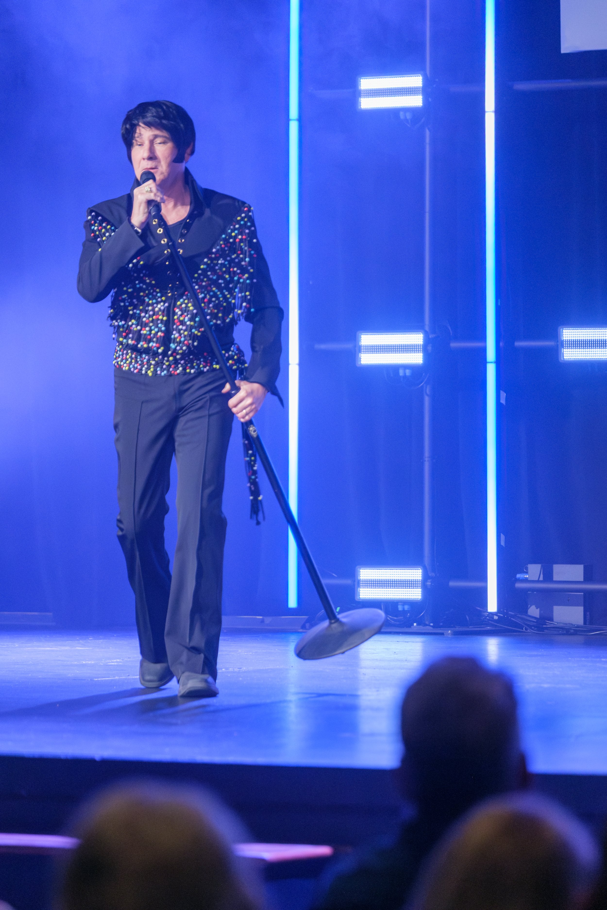 A person dressed as Elvis Presley performing on stage with a microphone in hand and a vintage microphone stand, wearing a black jumpsuit embellished with colorful studs, in front of vibrant blue and purple lighting.