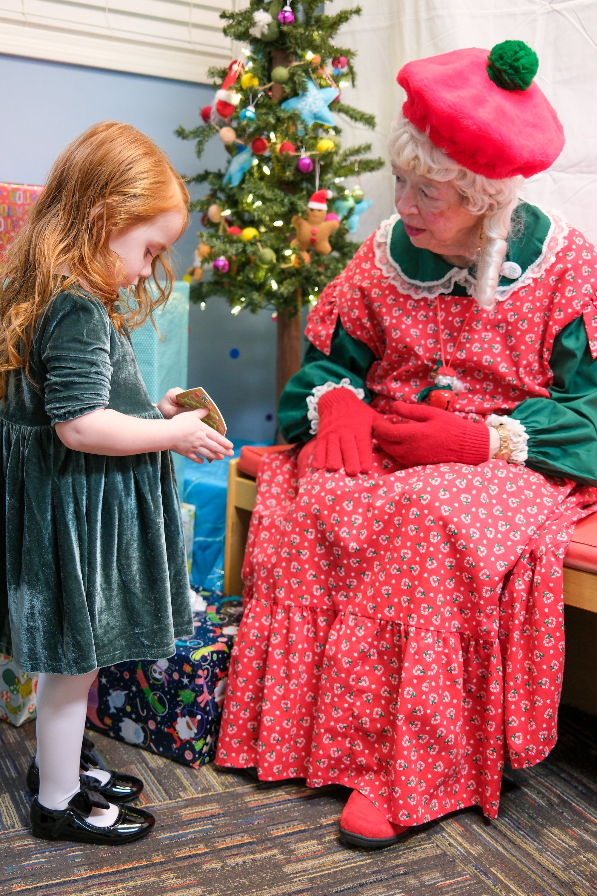 A young girl with long red hair gives a gift to Santa Claus, dressed as Mrs. Claus, during Christmas celebrations. A decorated Christmas tree is visible in the background.