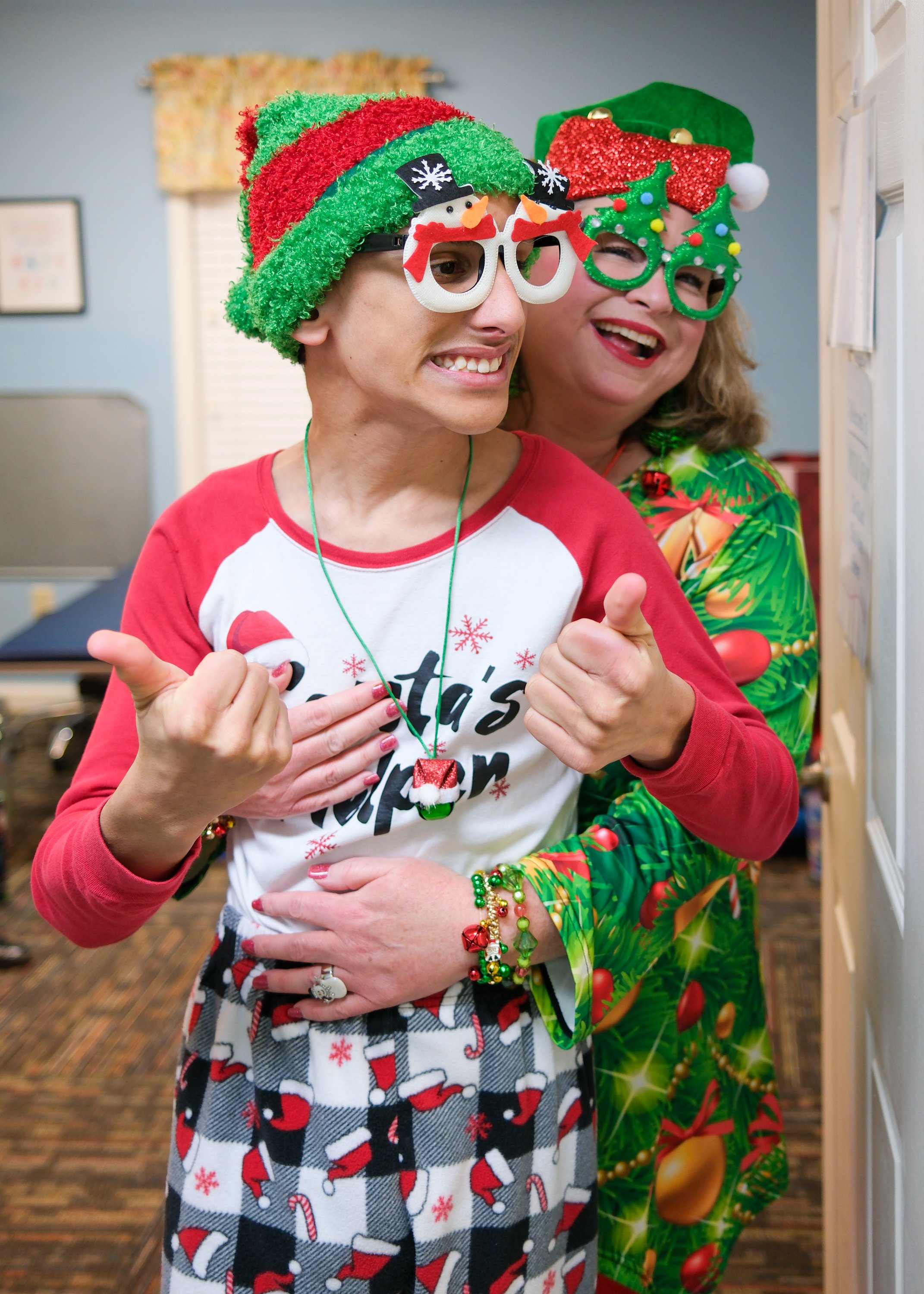 Two women celebrating Christmas in festive attire, wearing Christmas hats and glasses, with one in a Santa-themed shirt and the other in a holiday dress, happily standing together indoors.