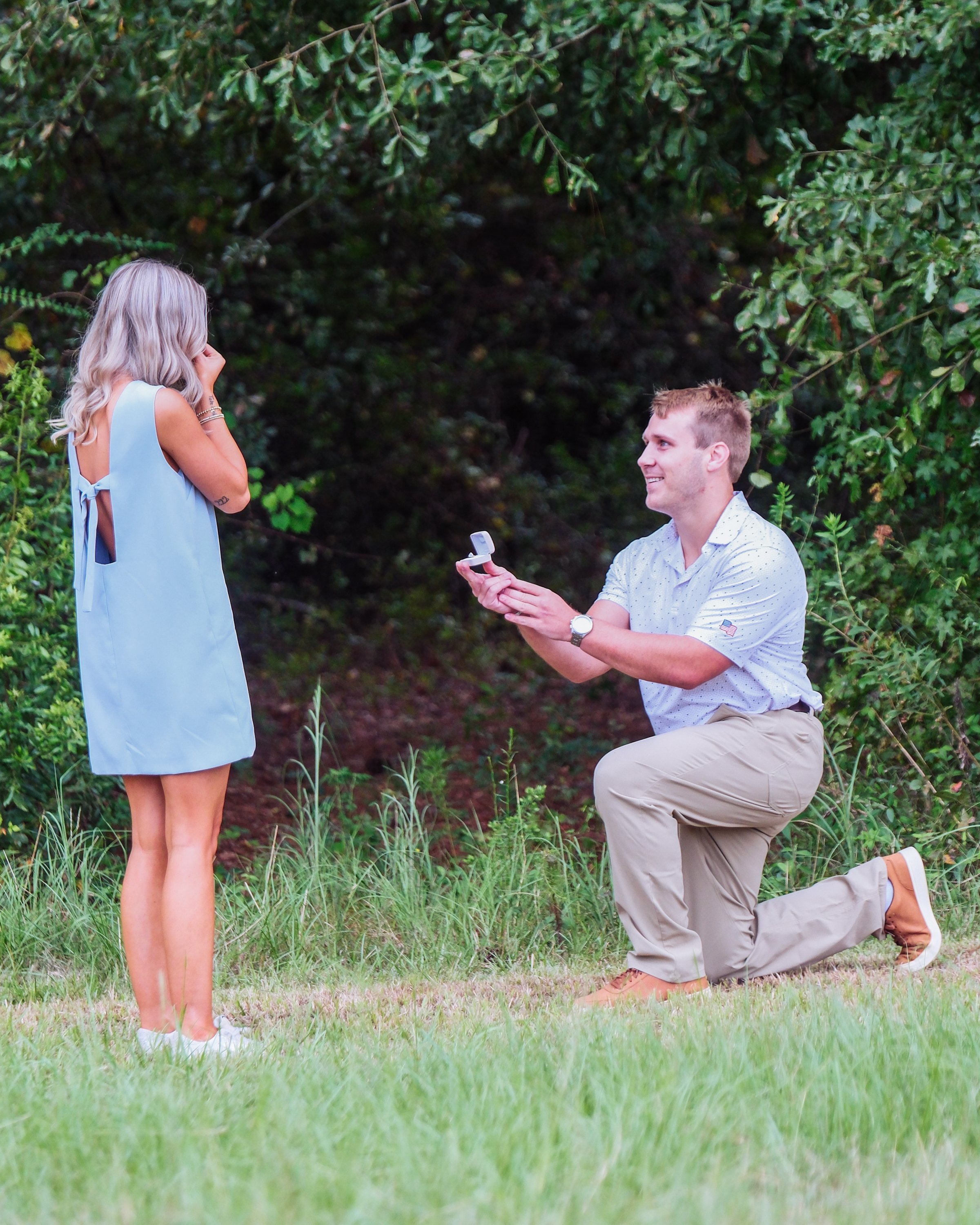 A man kneeling with one knee on the grass, proposing to a woman who appears surprised or emotional in a wooded outdoor setting.