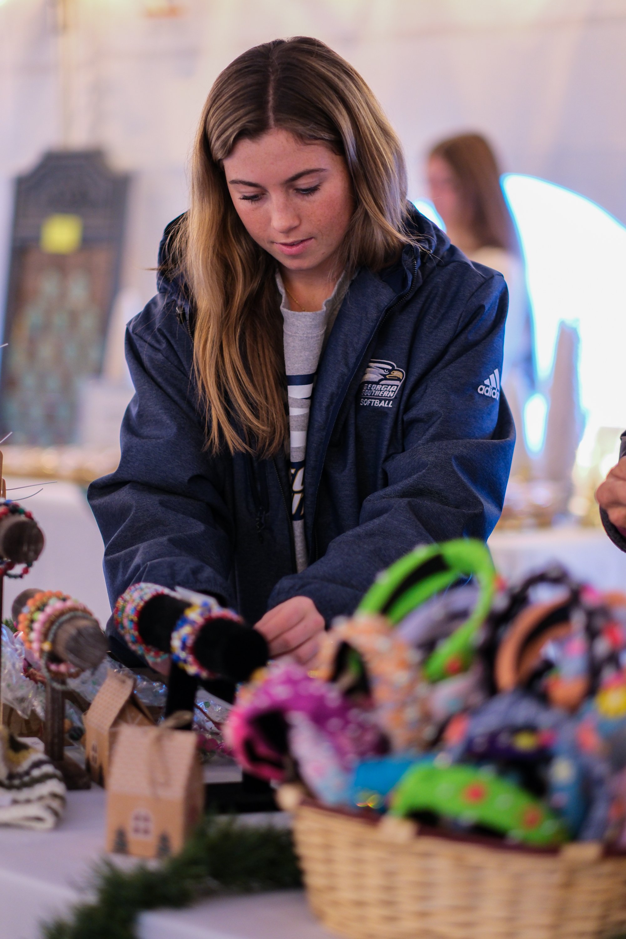 Young woman with light brown hair browsing colorful handmade accessories at a craft fair.