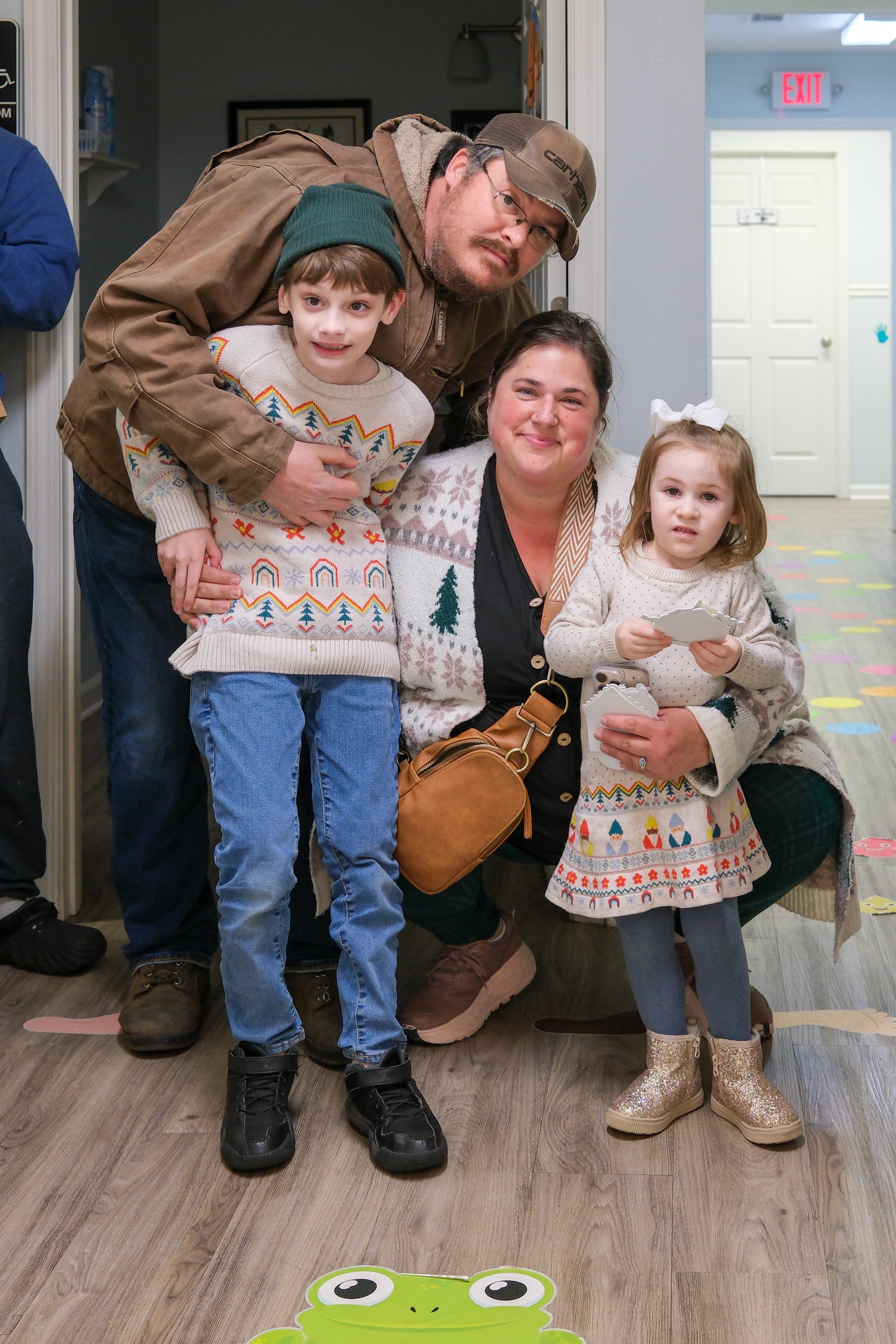Family group photo with two children, one girl and one boy, and two adults, inside a building with decorative floor stickers and a frog cutout on the floor.