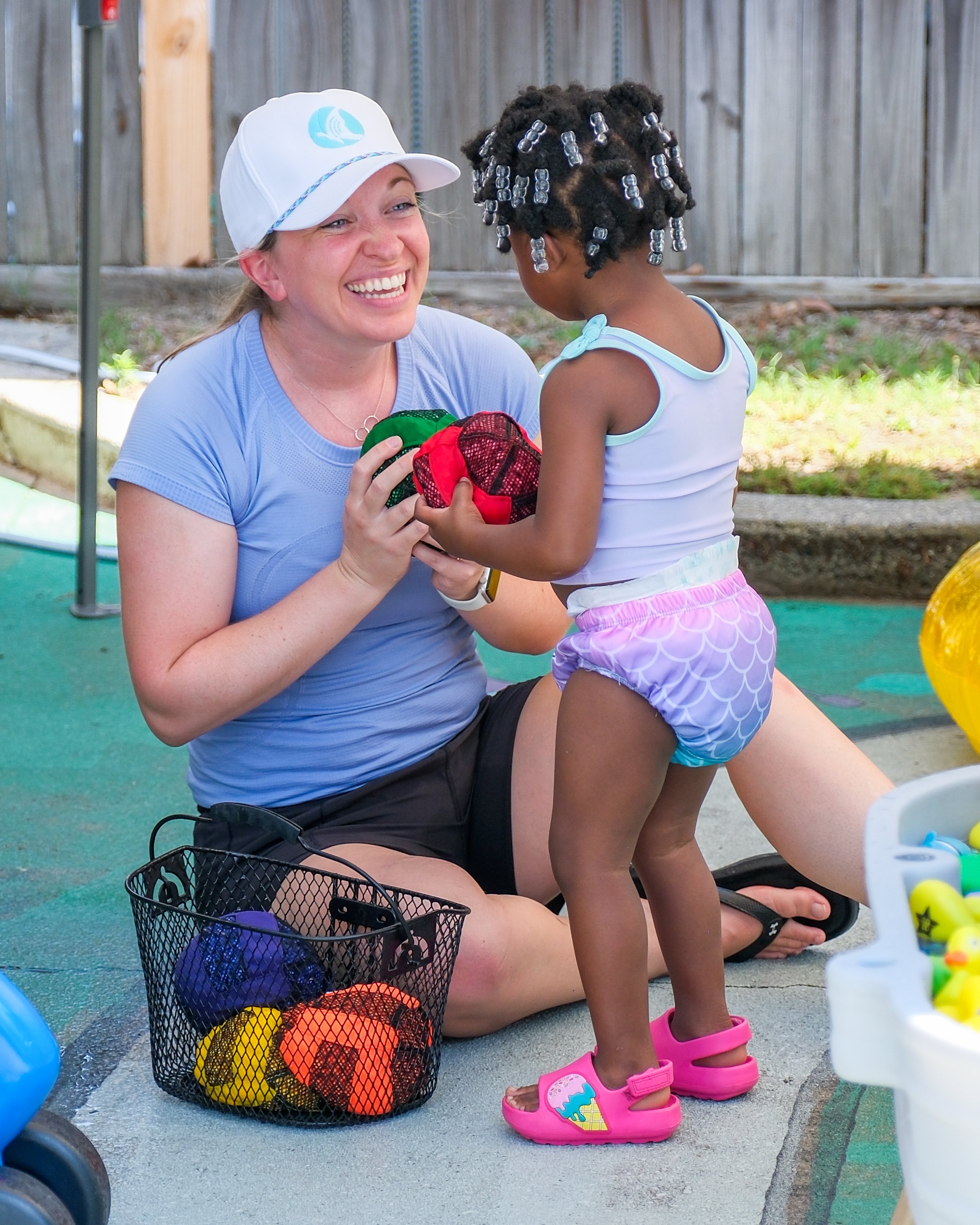 A woman with a baseball cap kneeling and smiling at a young girl, holding and exchanging small bags of toys or balls at an outdoor playground.