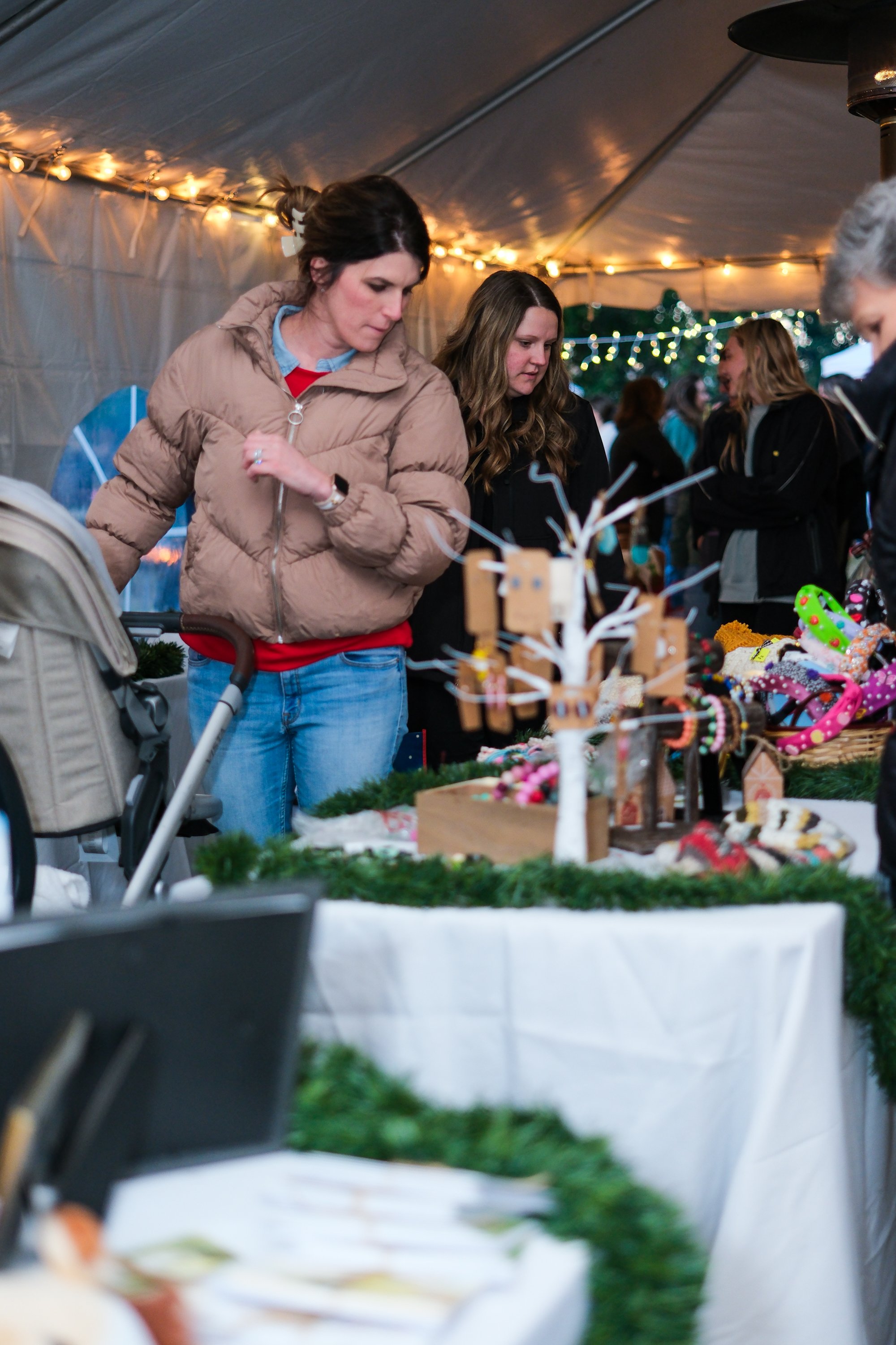 People shopping at a Christmas craft fair in a decorated tent with string lights, browsing holiday ornaments and accessories displayed on tables.