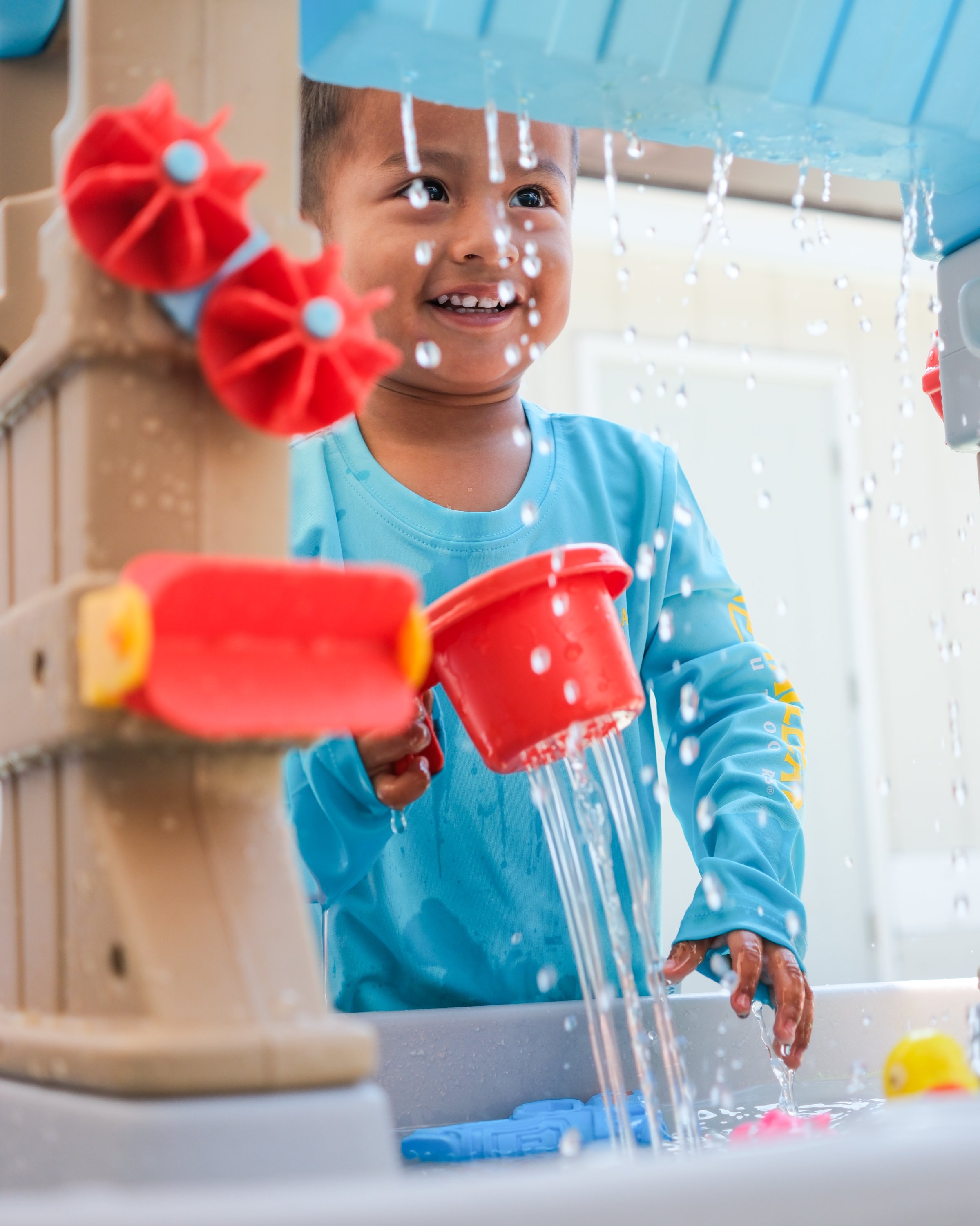 Young boy smiling and playing at a water table with red and yellow plastic toys, with water pouring from a blue structure above.