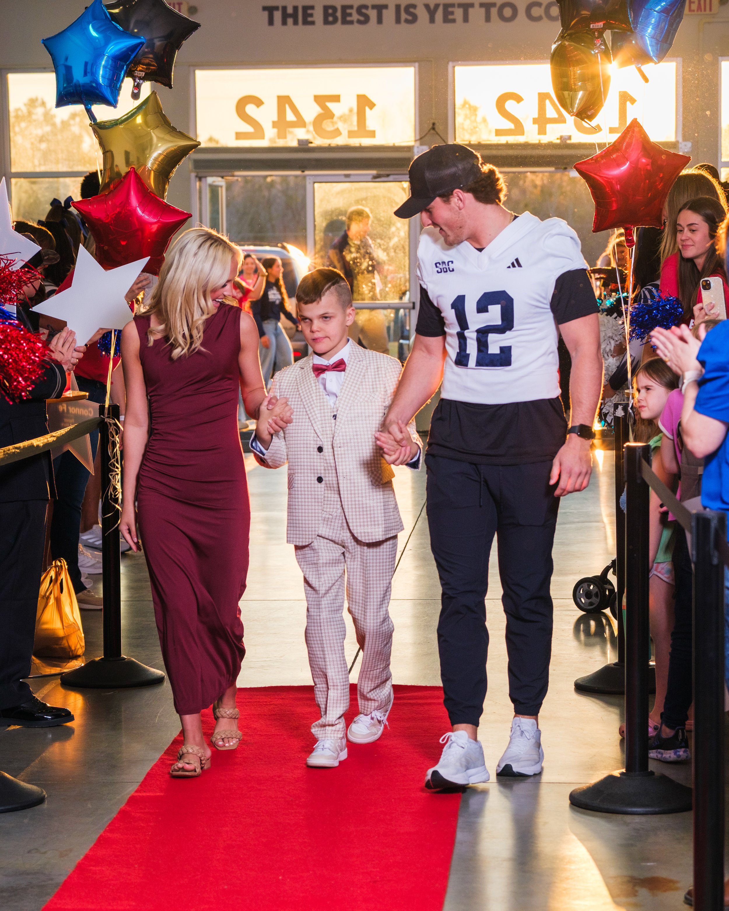 A young boy in a light-colored suit with a bow tie walks down a red carpet holding hands with a woman in a burgundy dress and a man in a white football jersey with the number 12. They are at an indoor event with balloons and other children and adults