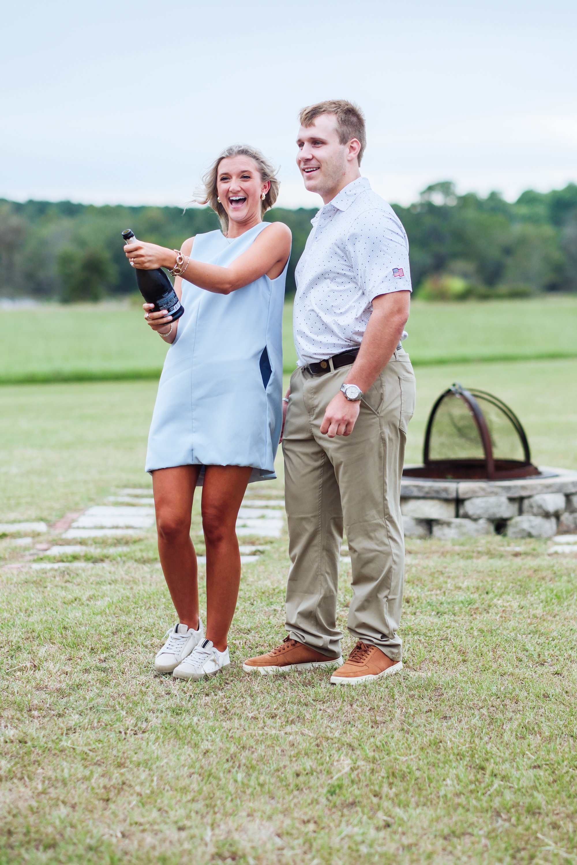 A young woman and man smiling and laughing outdoors, the woman holding a bottle of champagne, on a grassy area with a fire pit in the background.