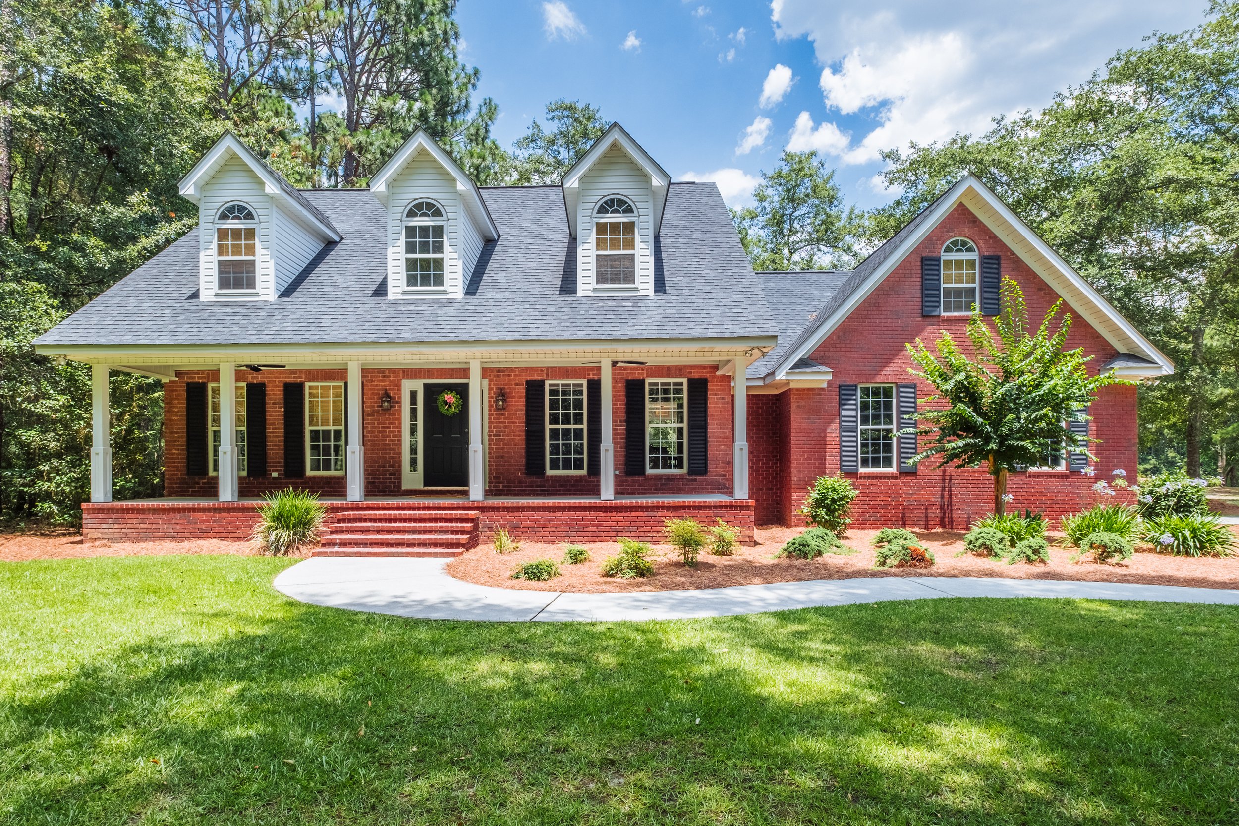 A two-story brick house with three dormer windows, a covered front porch, and black shutters, surrounded by green grass, plants, and trees under a blue sky with scattered clouds.