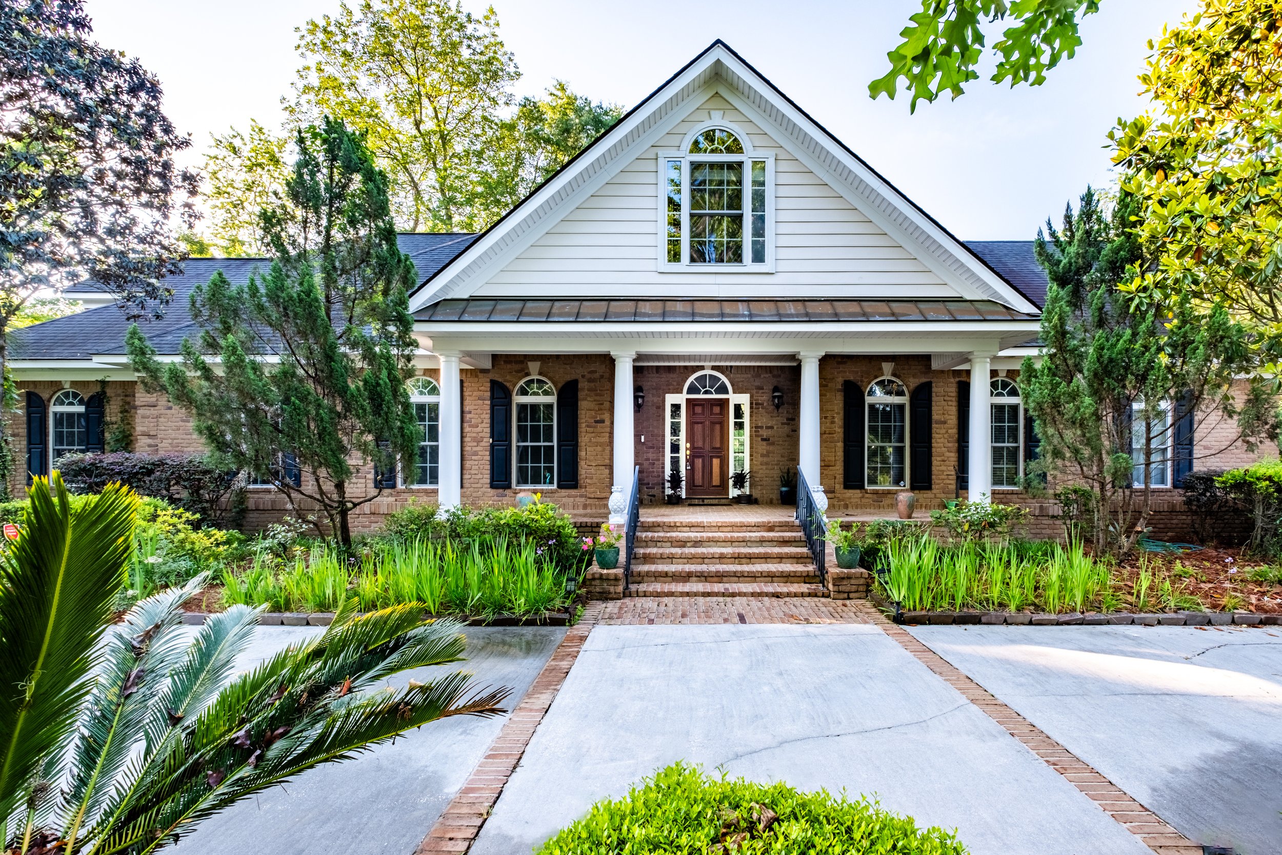 Front view of a house with brick exterior, white columns, and a gable roof surrounded by greenery and trees.
