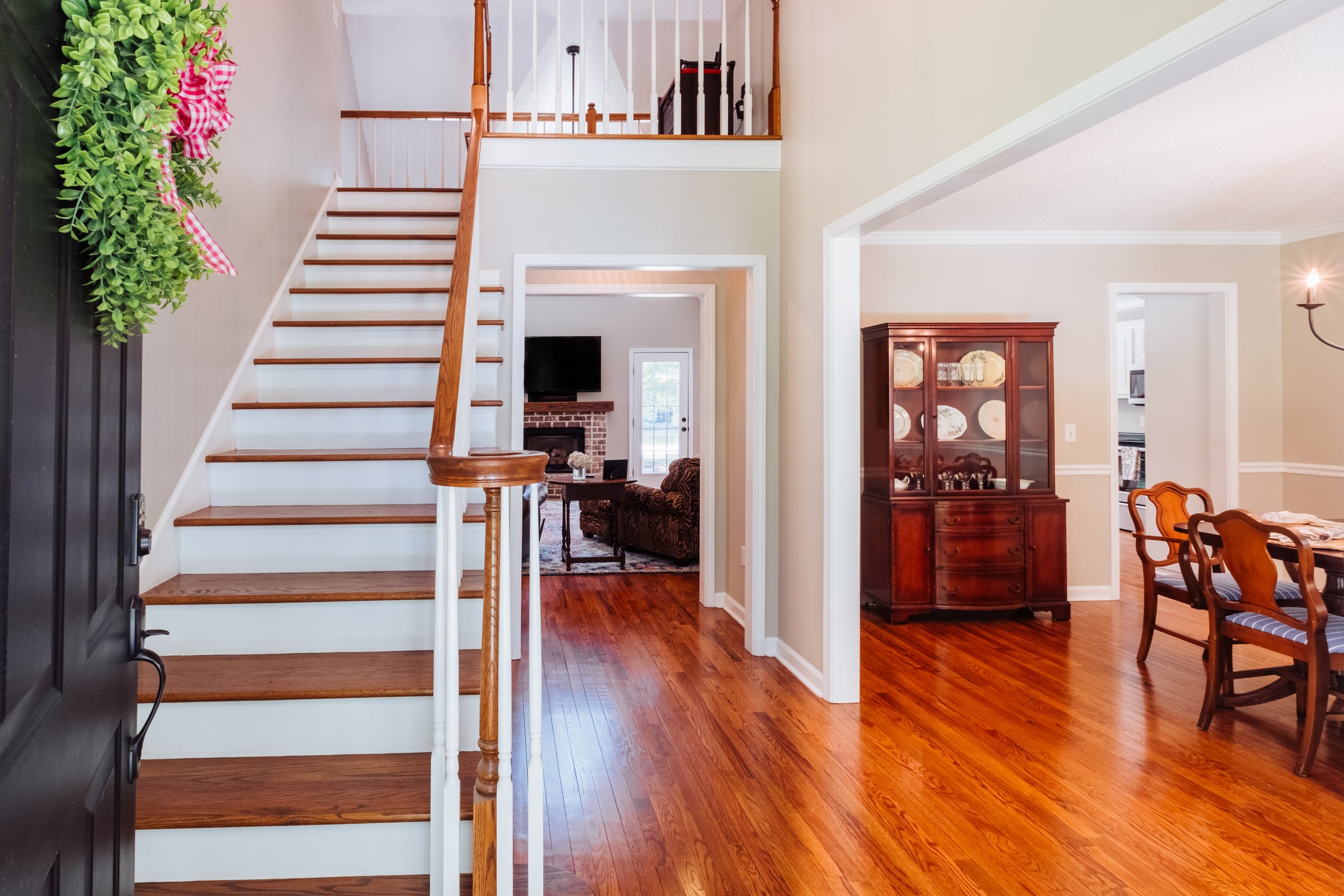 View of a home interior showing wooden stairs, a dining area with chairs and a hutch, and a living room with a fireplace and TV.
