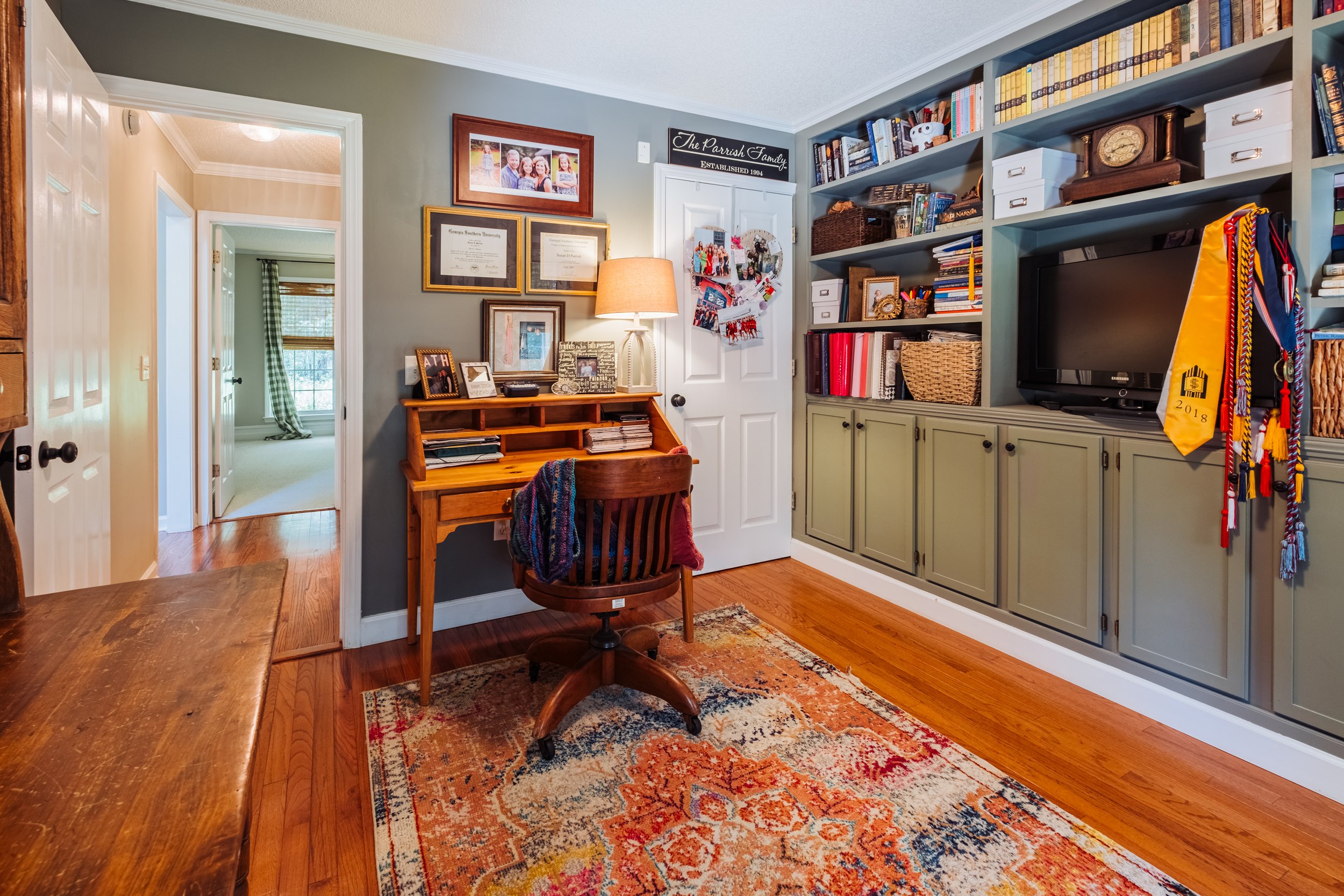 Home office or study room with a wooden desk, office chair, built-in bookshelf with books and decor, and a wall with framed photos and certificates.