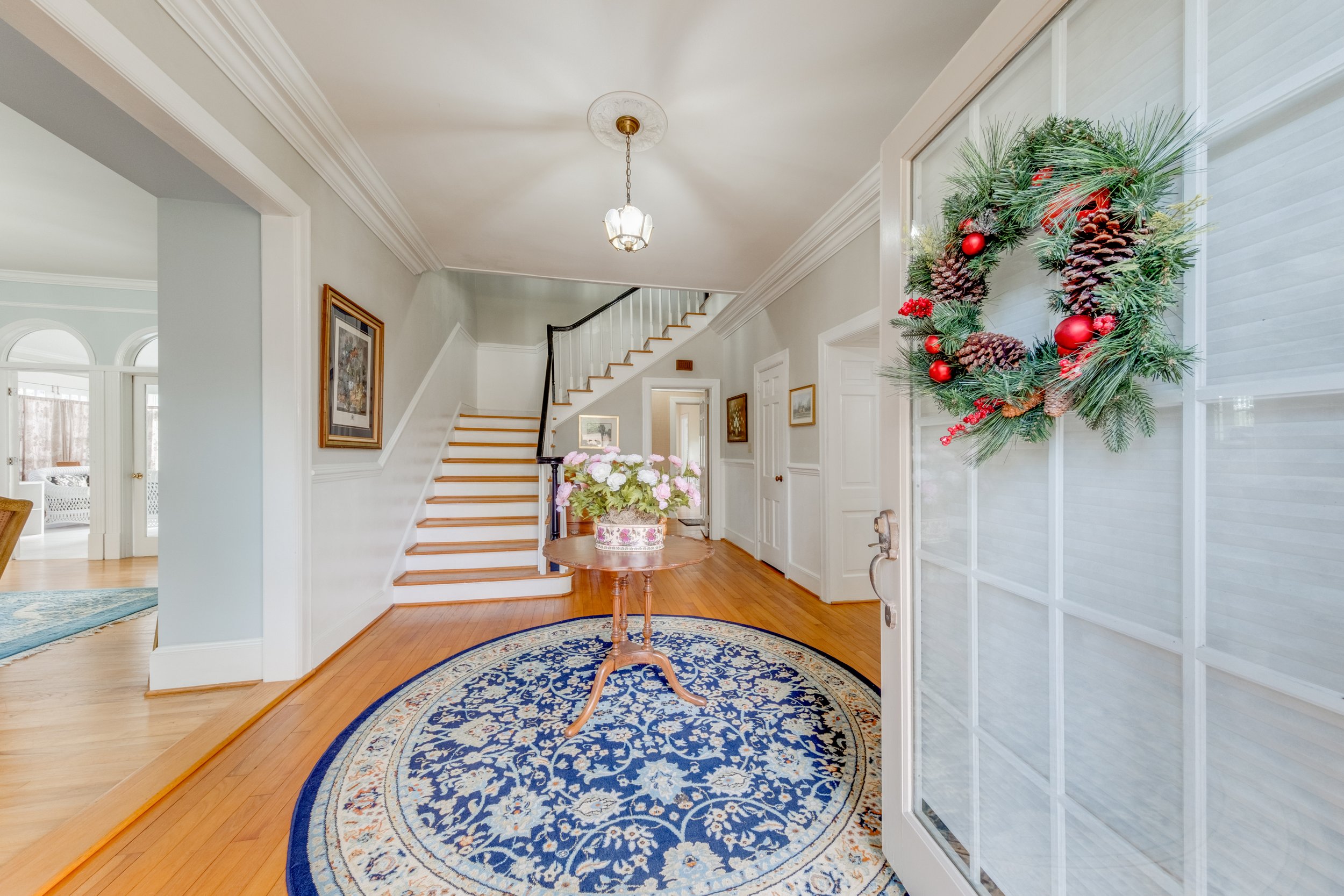 Entryway of a house decorated for Christmas with a wreath on the door, a round blue and cream rug, a small wooden table with a flower arrangement, and a staircase in the background.