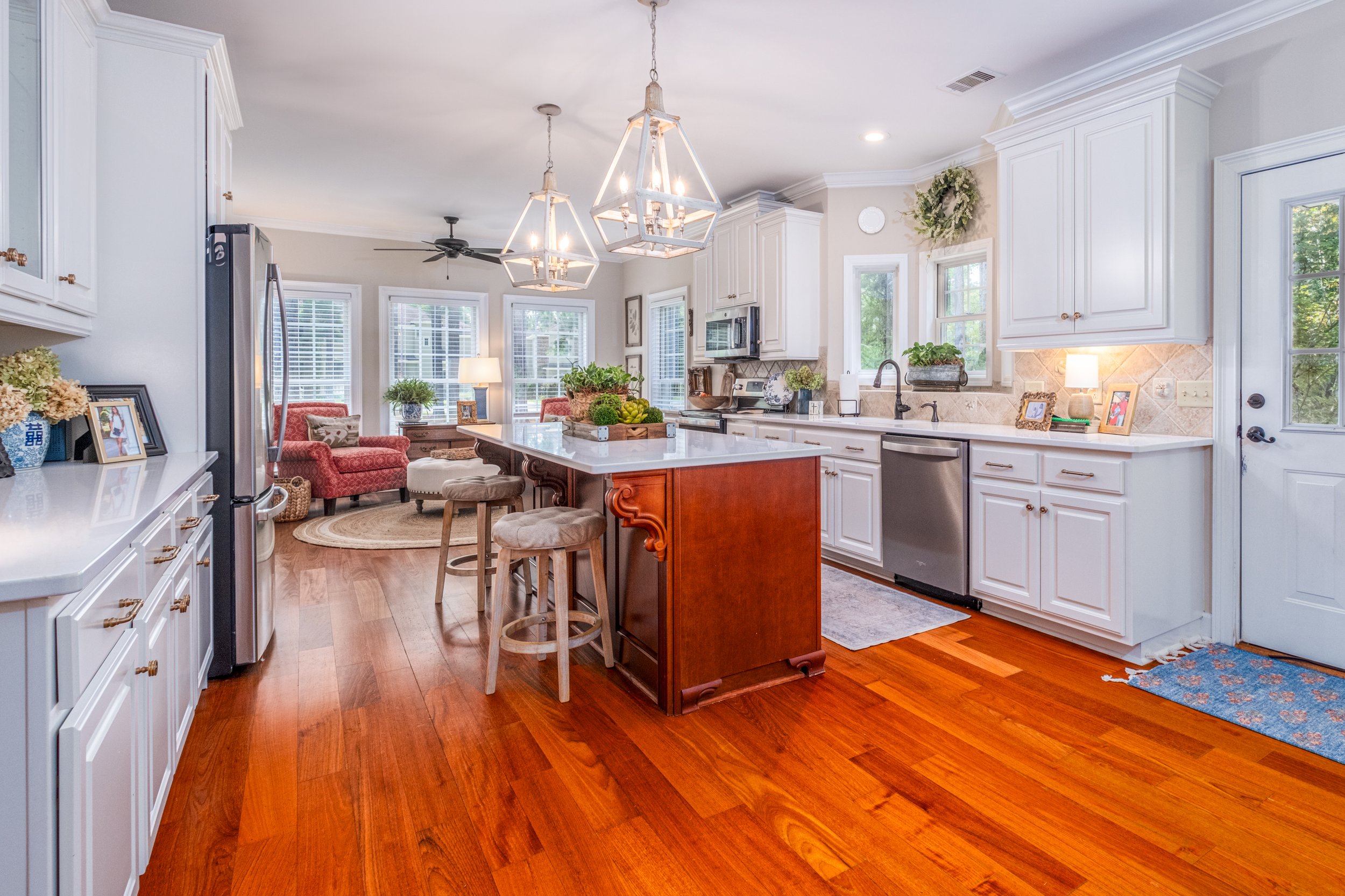 Bright and spacious kitchen with white cabinetry, wooden island with countertop, pendant lights, stainless steel appliances, hardwood floor, and a view into a cozy sitting area with red furniture and large windows.