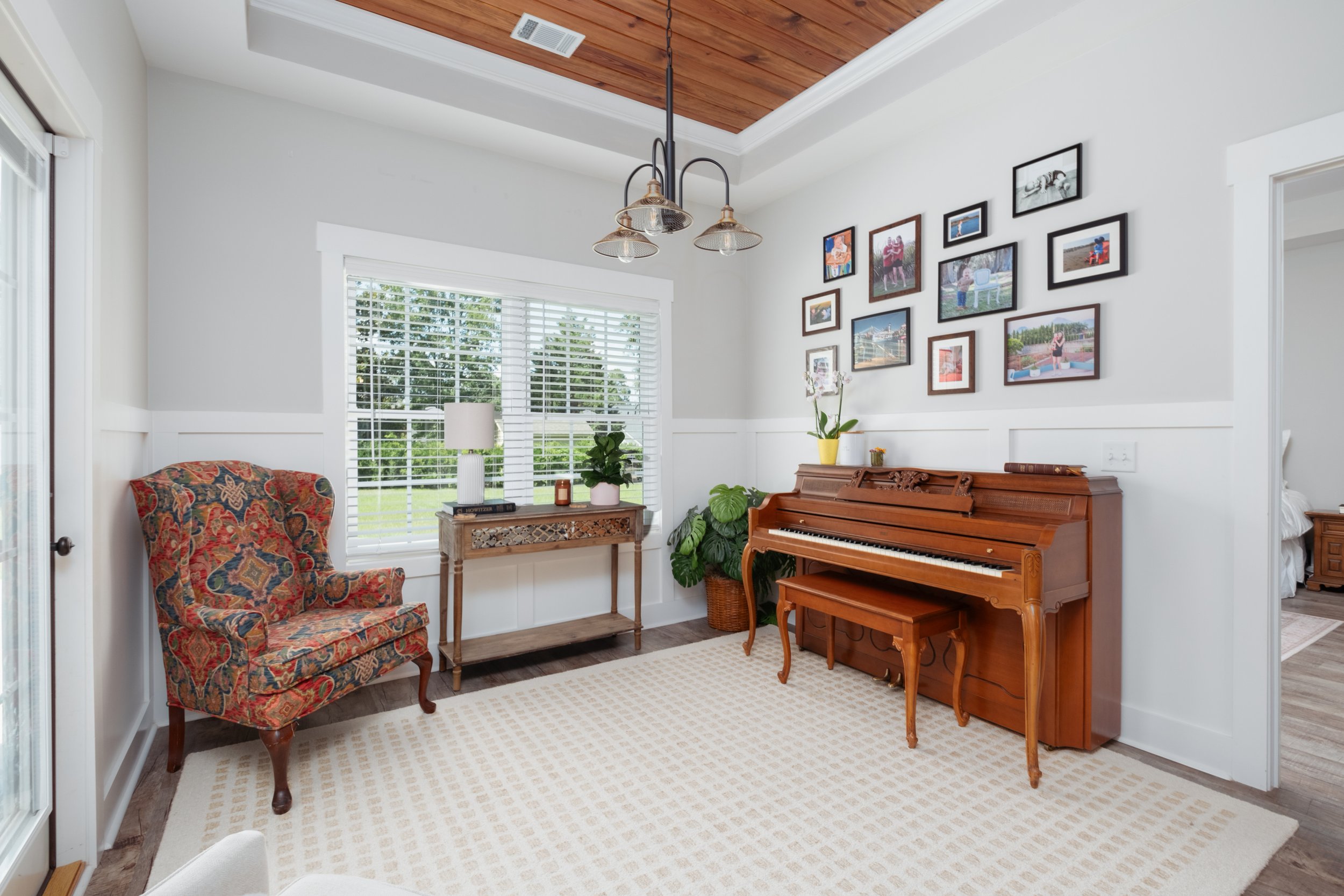 Living room with a wooden upright piano, framed photos on the wall, an armchair with a floral pattern, a window with blinds, and a wall-mounted light fixture.