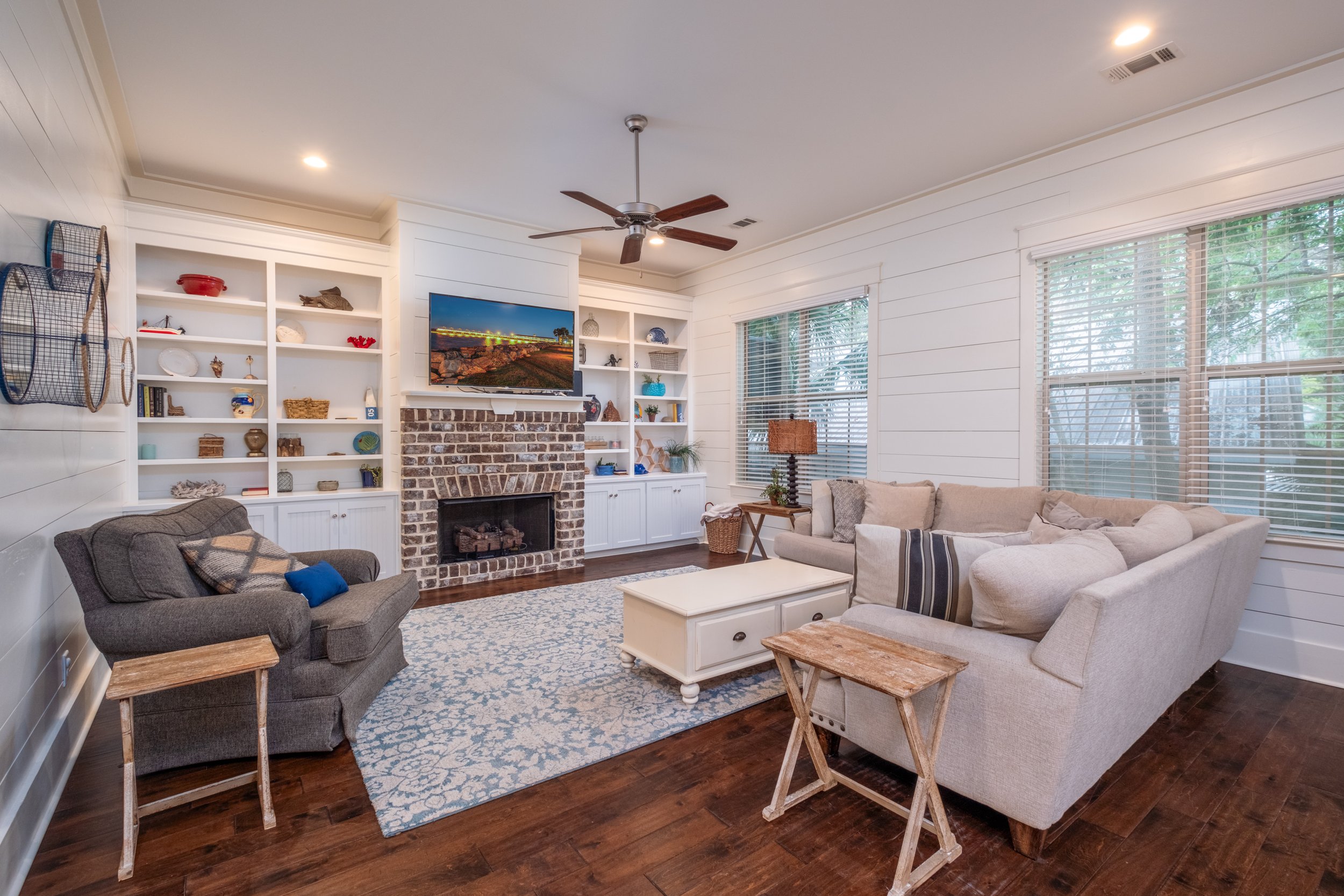 Living room with white paneled walls, a brick fireplace, and a mounted flat-screen TV. Furniture includes a beige sofa with pillows, a gray armchair with a pillow, and small wooden side tables. Large windows with blinds let in natural light, and book