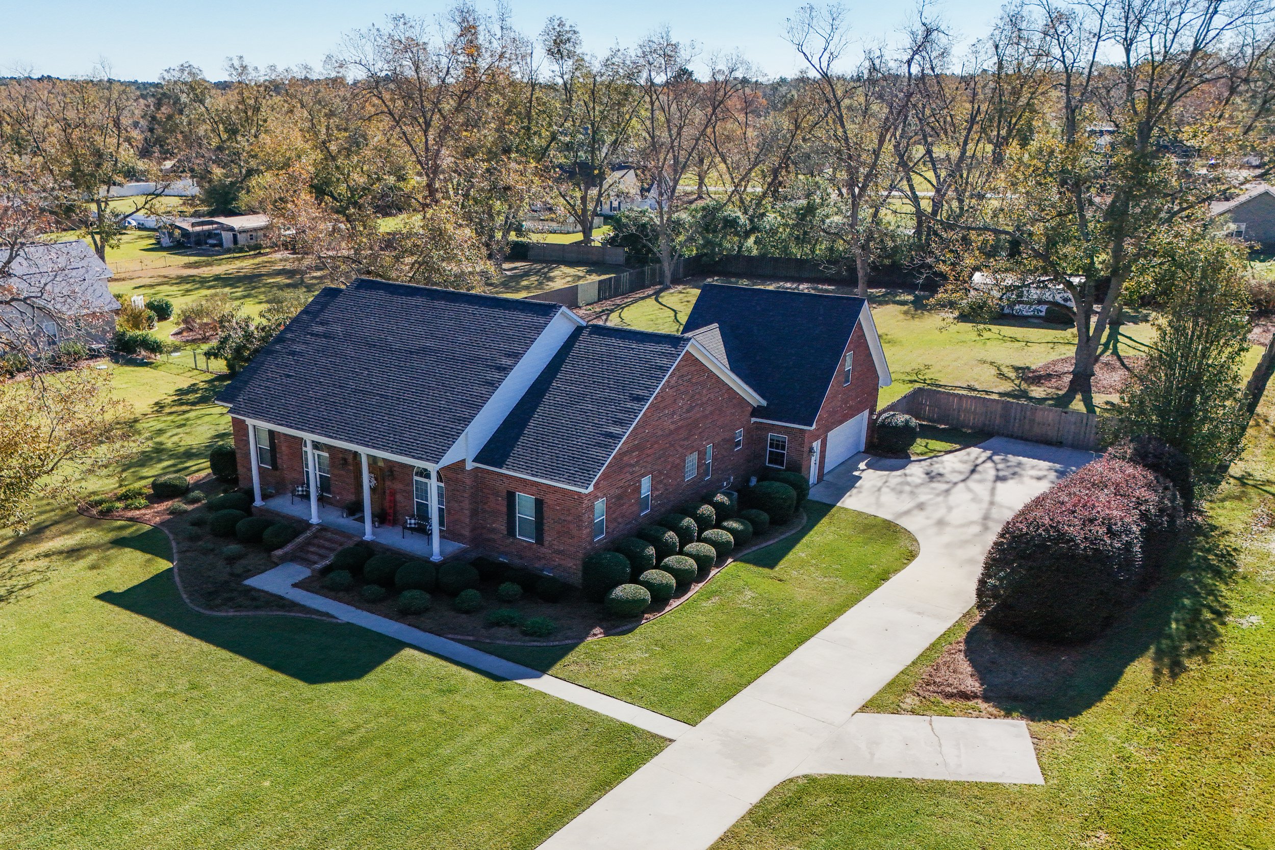 Aerial view of a brick house with a front porch and well-maintained lawn, surrounded by trees and a concrete driveway leading to a garage, in a suburban neighborhood.