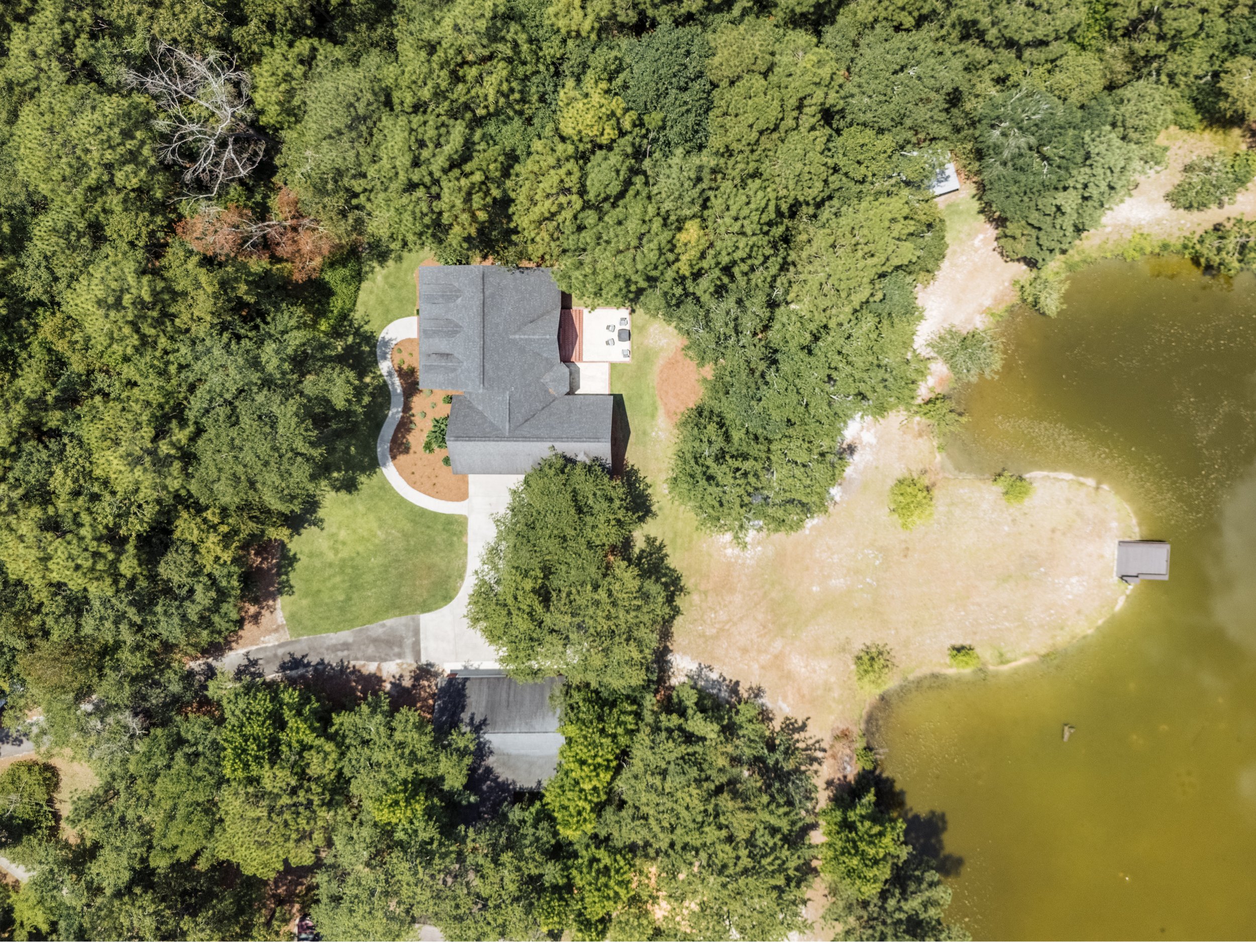 Aerial view of a house surrounded by trees, near a pond, with a driveway and lawn.
