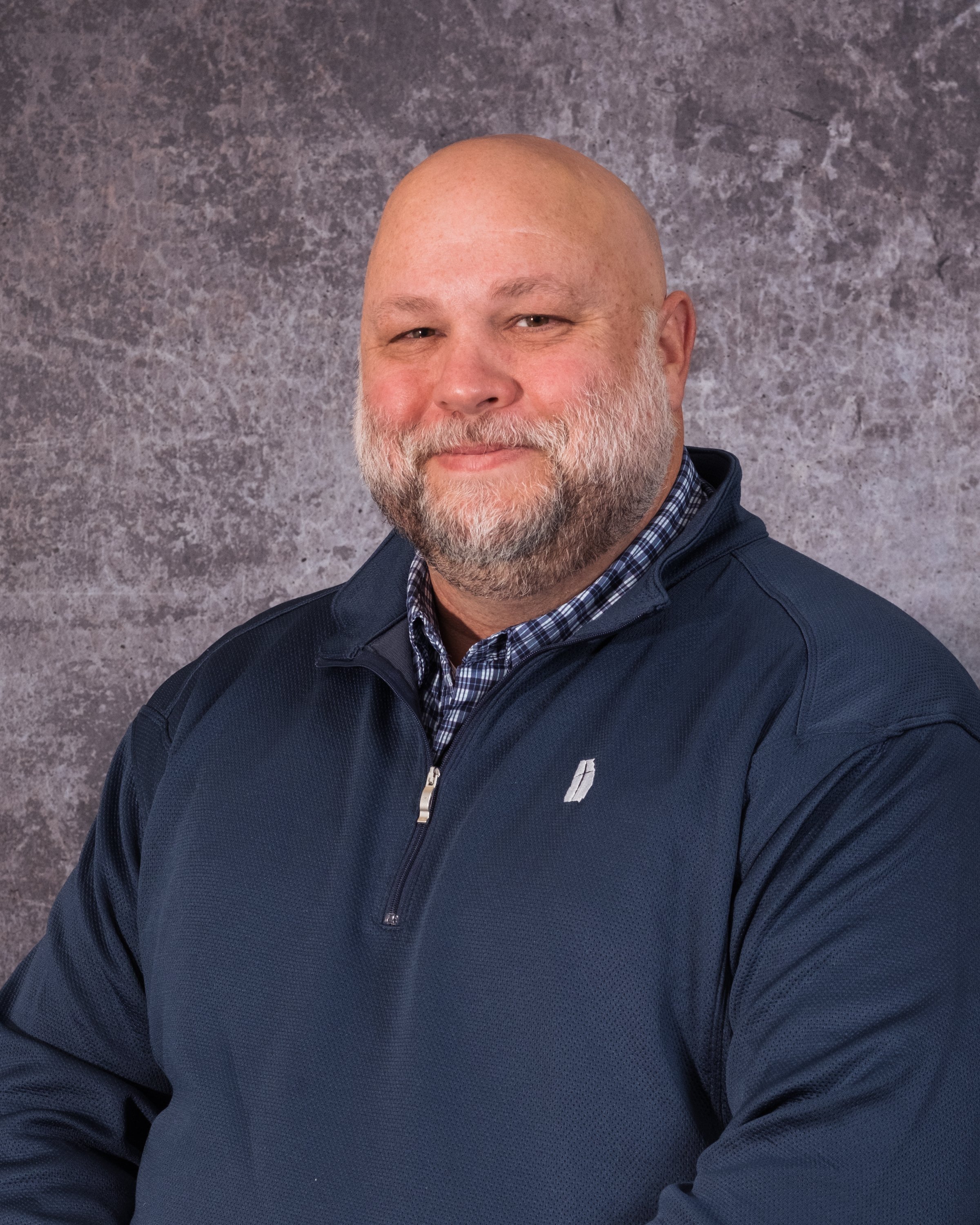A middle-aged man with a bald head, beard, and mustache, wearing a navy blue zip-up jacket over a checkered shirt, smiling softly, in front of a textured gray background.