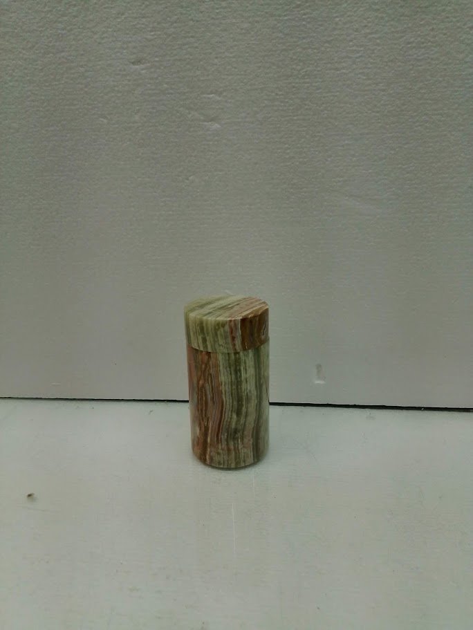 A small, cylindrical object with a rounded top, made of multicolored stone or marble, placed on a white surface against a light-colored wall.