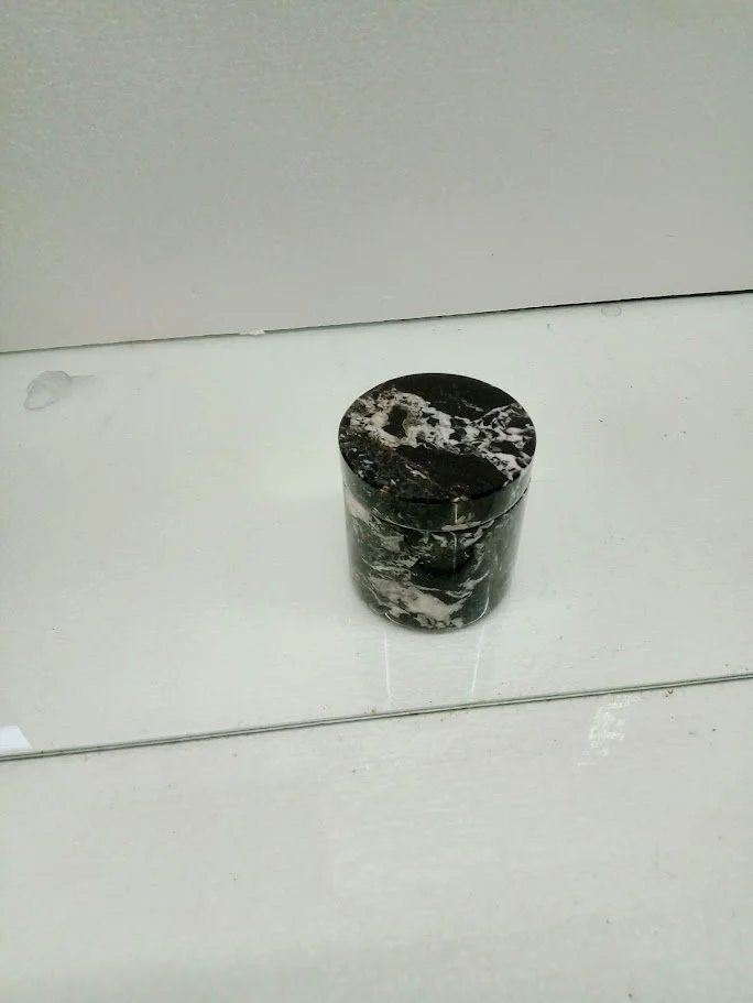 A small black and white marble cylindrical container with a tight-fitting lid, placed on a white surface in front of a mirror.