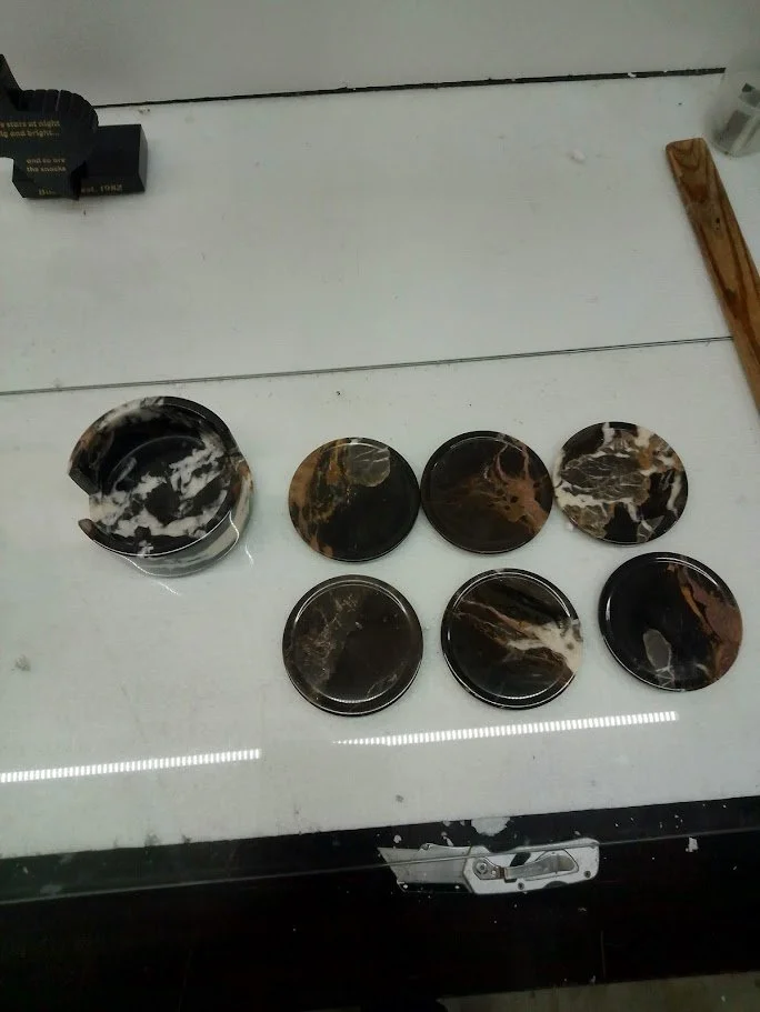 Collection of seven round black and brown marble-patterned coasters on a white table, with a partially visible black object and wooden support in the background.