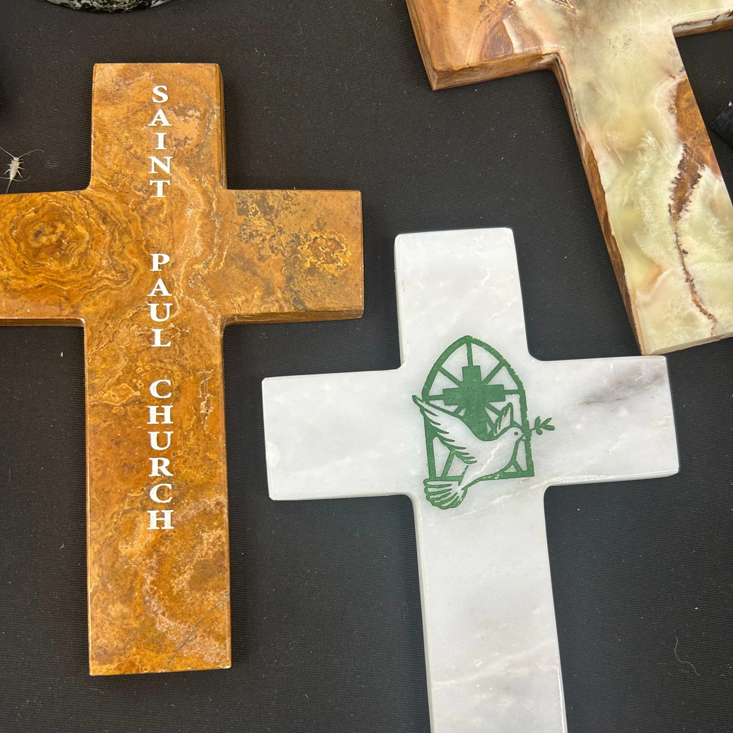 Three crosses made of different stones on a black surface. The first cross is rust-colored with the text 'Saint Paul Church' written vertically in white. The second cross is white with a green dove and a cross symbol engraved on it. The third cross, partially visible, has a marbled pattern with shades of beige and brown.
