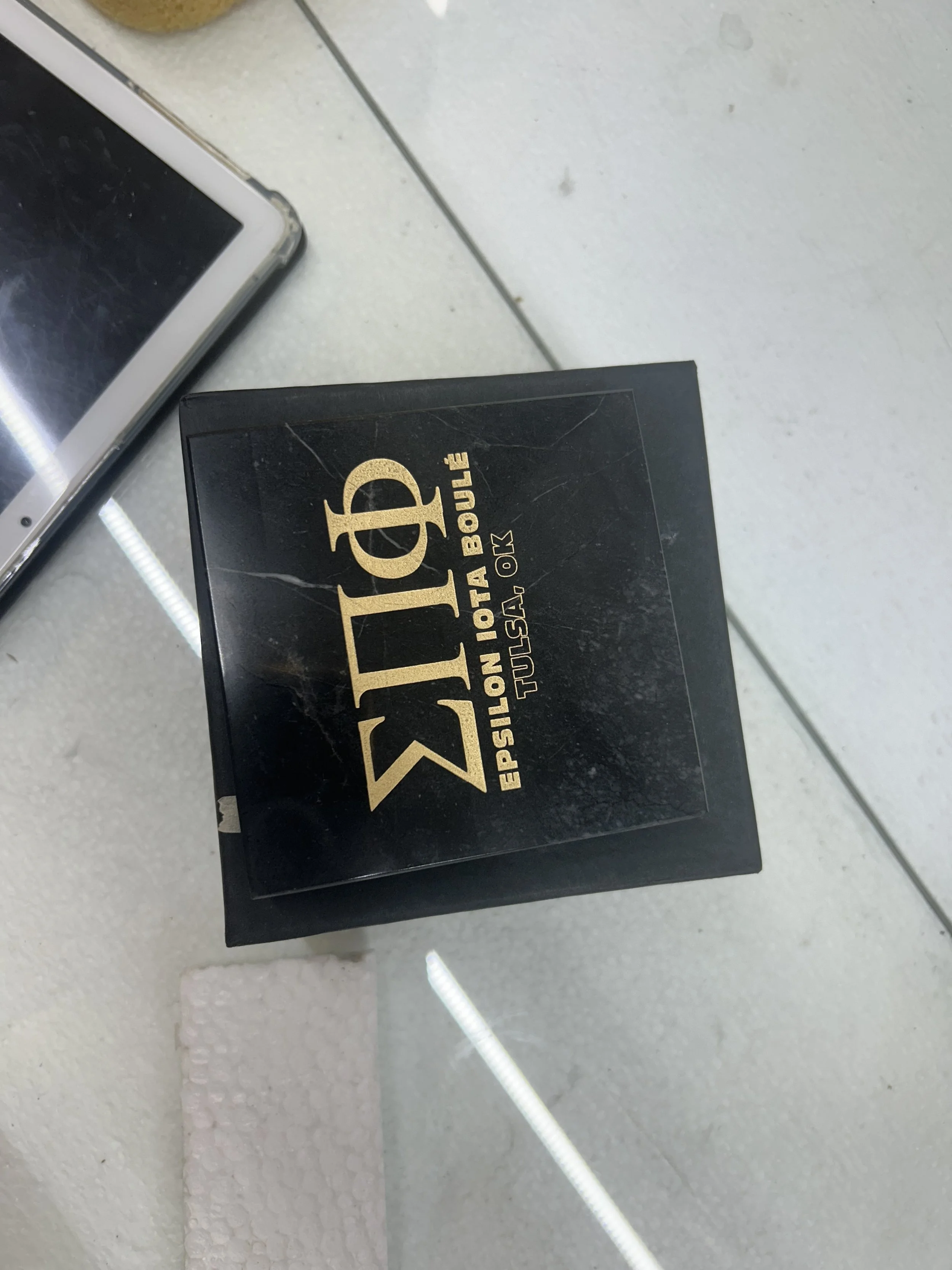 Black box with gold text labeled 'S.T.D. Epsilon Ioleta Boule Tulsa USA OK' placed on a white surface, with part of a smartphone visible nearby.