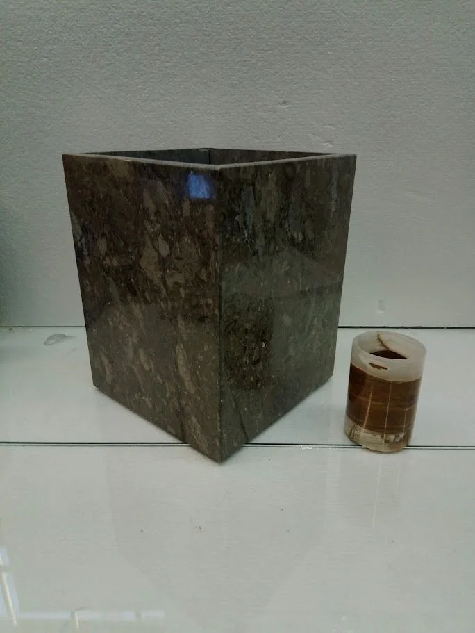 A dark brown square-shaped marble planter and a small beige container with a dark liquid on a white wooden surface.