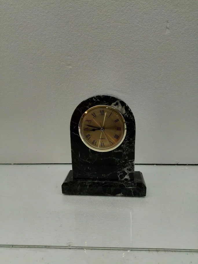 A black marble desk clock with gold face and Roman numerals, placed on a white surface against a plain wall.