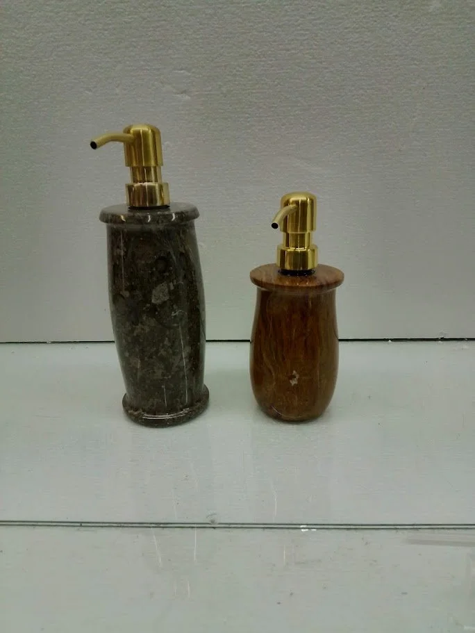 Two soap dispensers with gold pumps, one made of dark marble and the other of wood, placed on a white surface against a white wall.