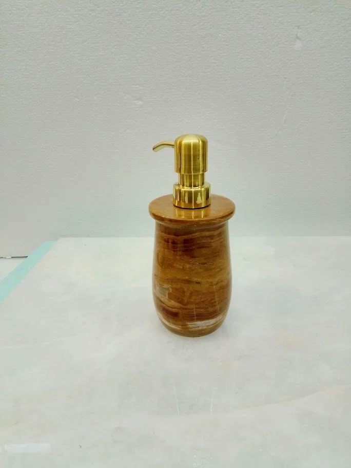 A soap dispenser with a gold pump on a wooden stand, placed on a white surface against a plain wall.