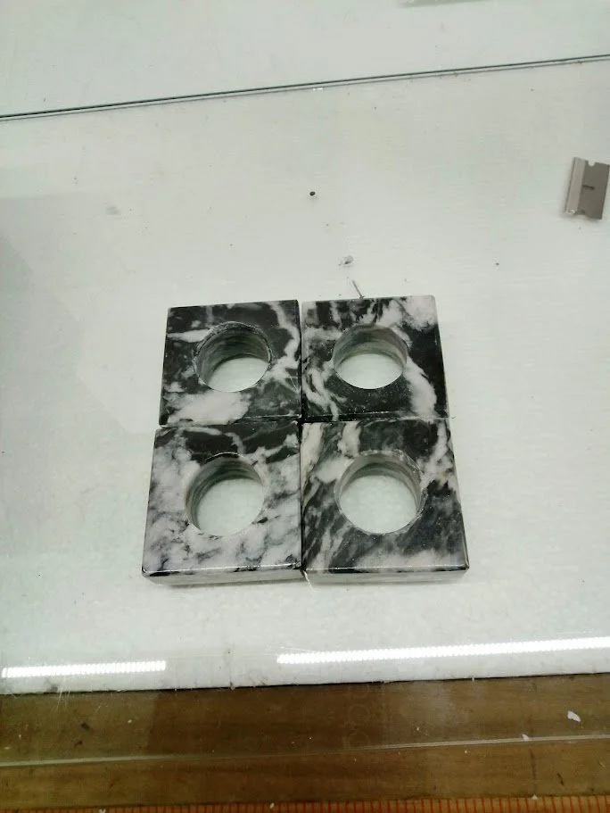 Four black and white marble-patterned tiles arranged in a square on a marble countertop. Each tile has a circular hole in the center.