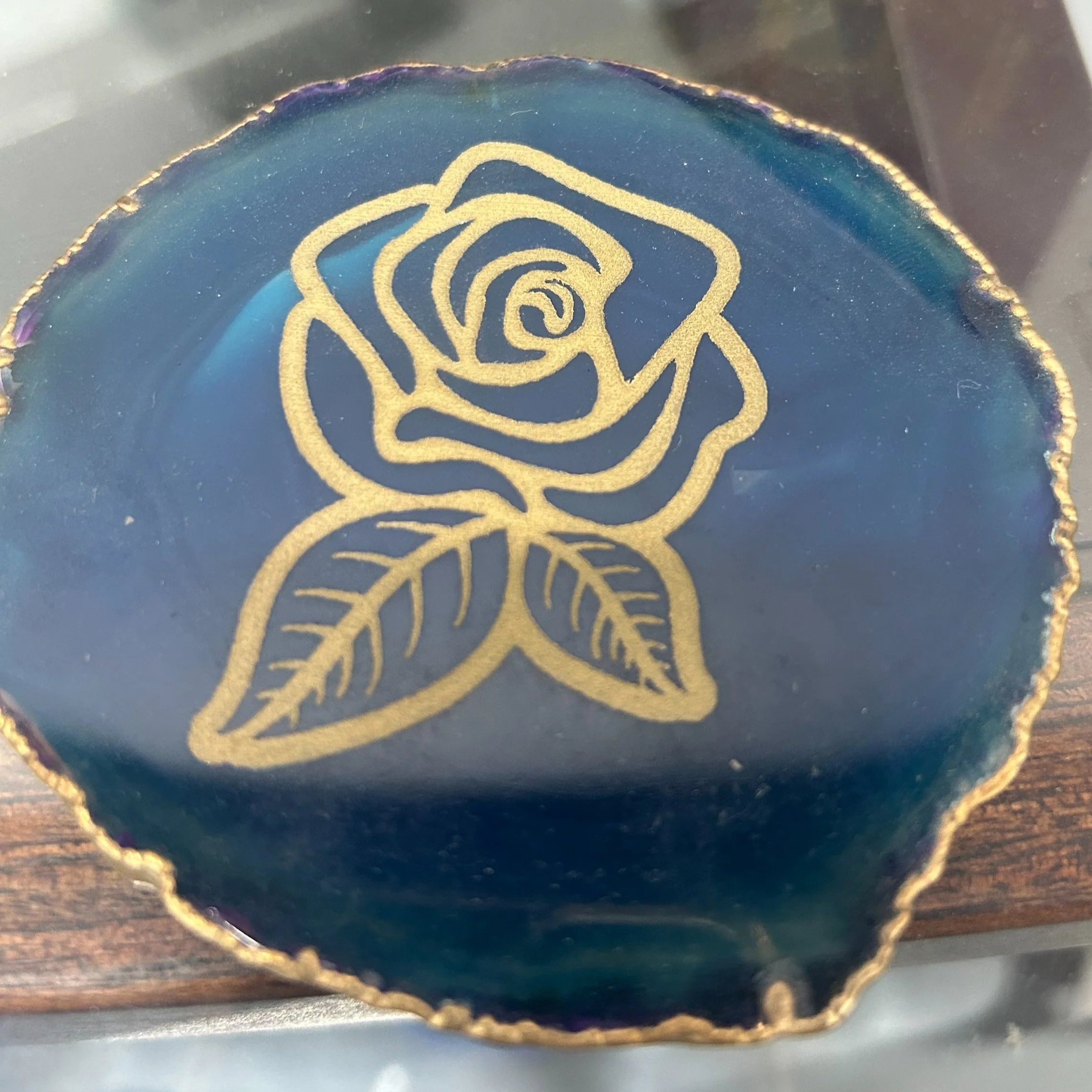 A decorative blue agate stone with a gold outline of a rose and leaves engraved on it.
